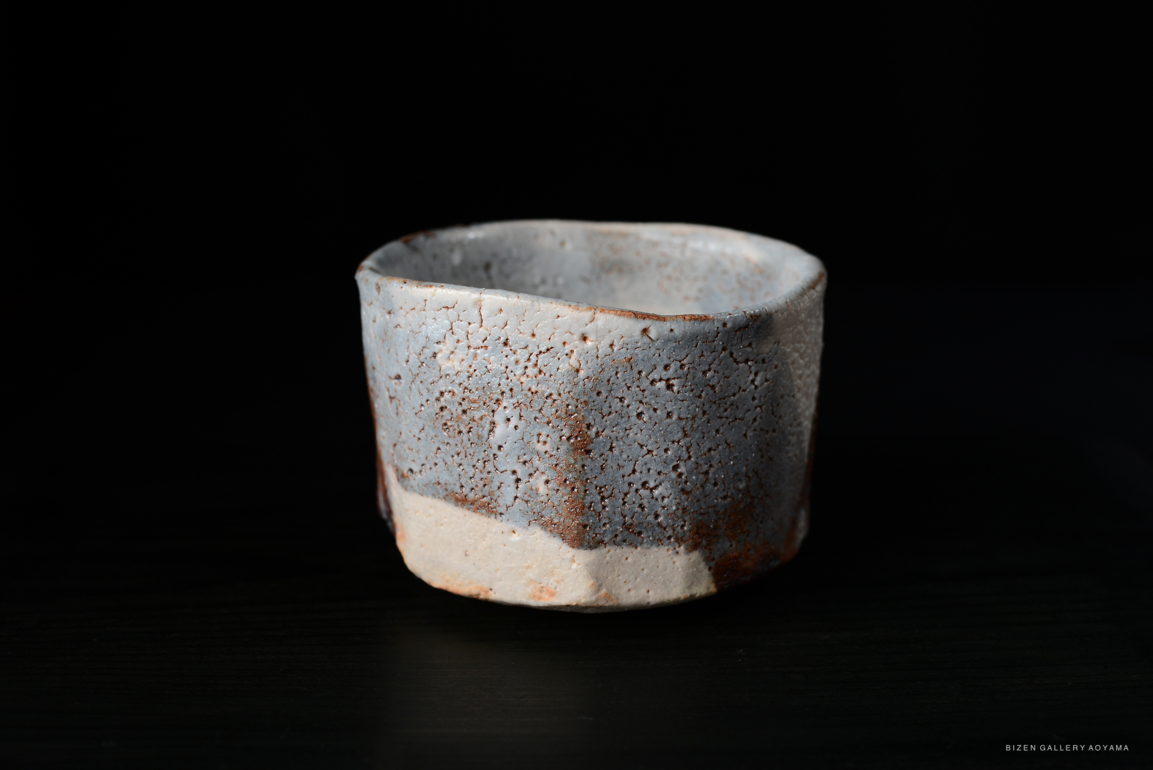 A Nezumi Shino Chawan tea bowl featuring pale blue-grey glaze with a speckled texture and rusty accents, displayed against a black background.