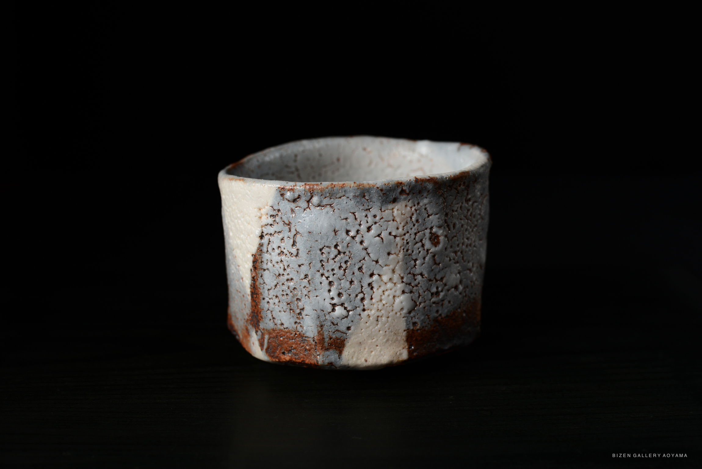 A rustic ceramic tea bowl featuring a textured, speckled surface with pale blue-grey hues and earthy rust details, set against a dark background.