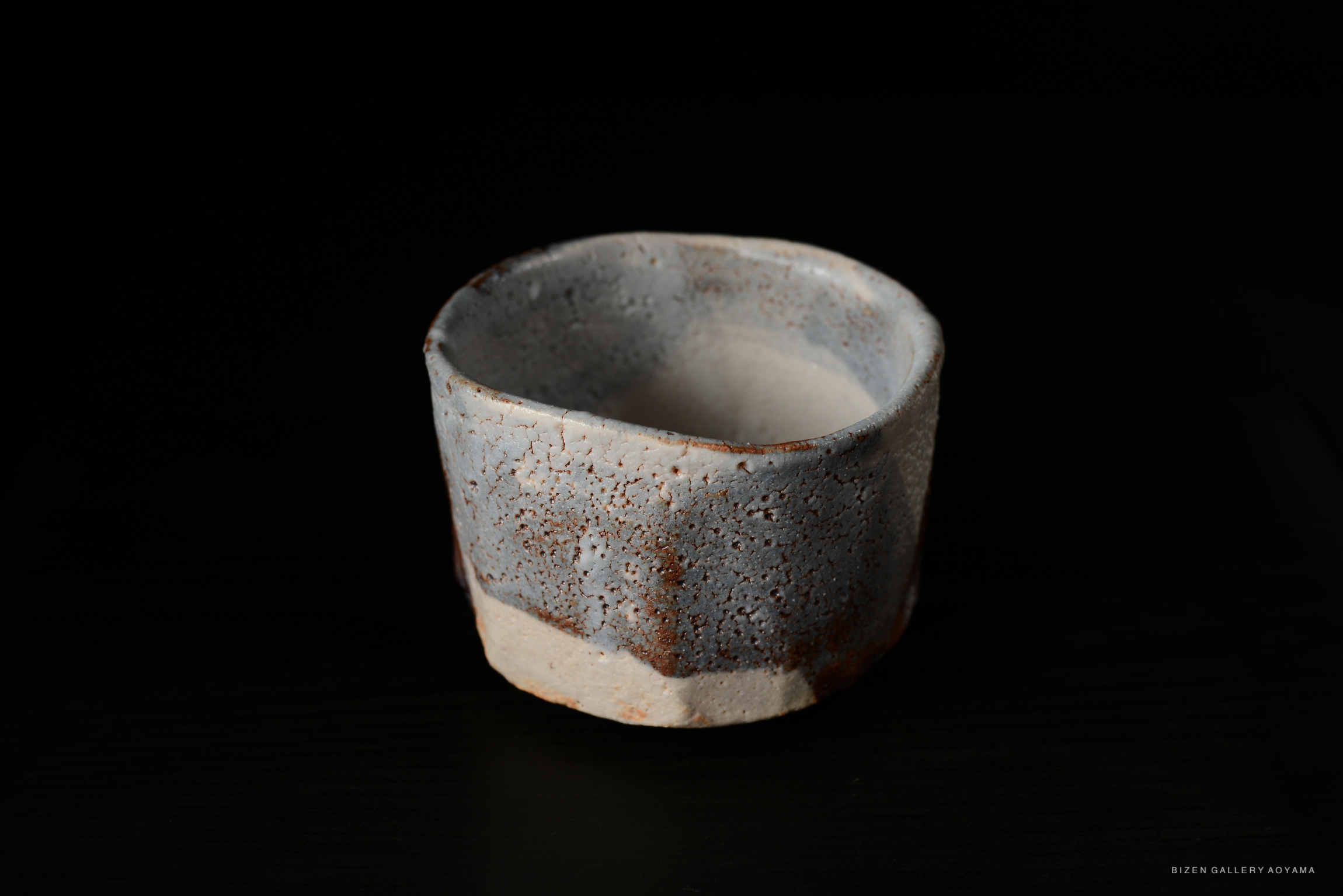 A Shino Chawan tea bowl with a pale blue-grey color and rustic finish, highlighting its unique shape and texture against a dark background.