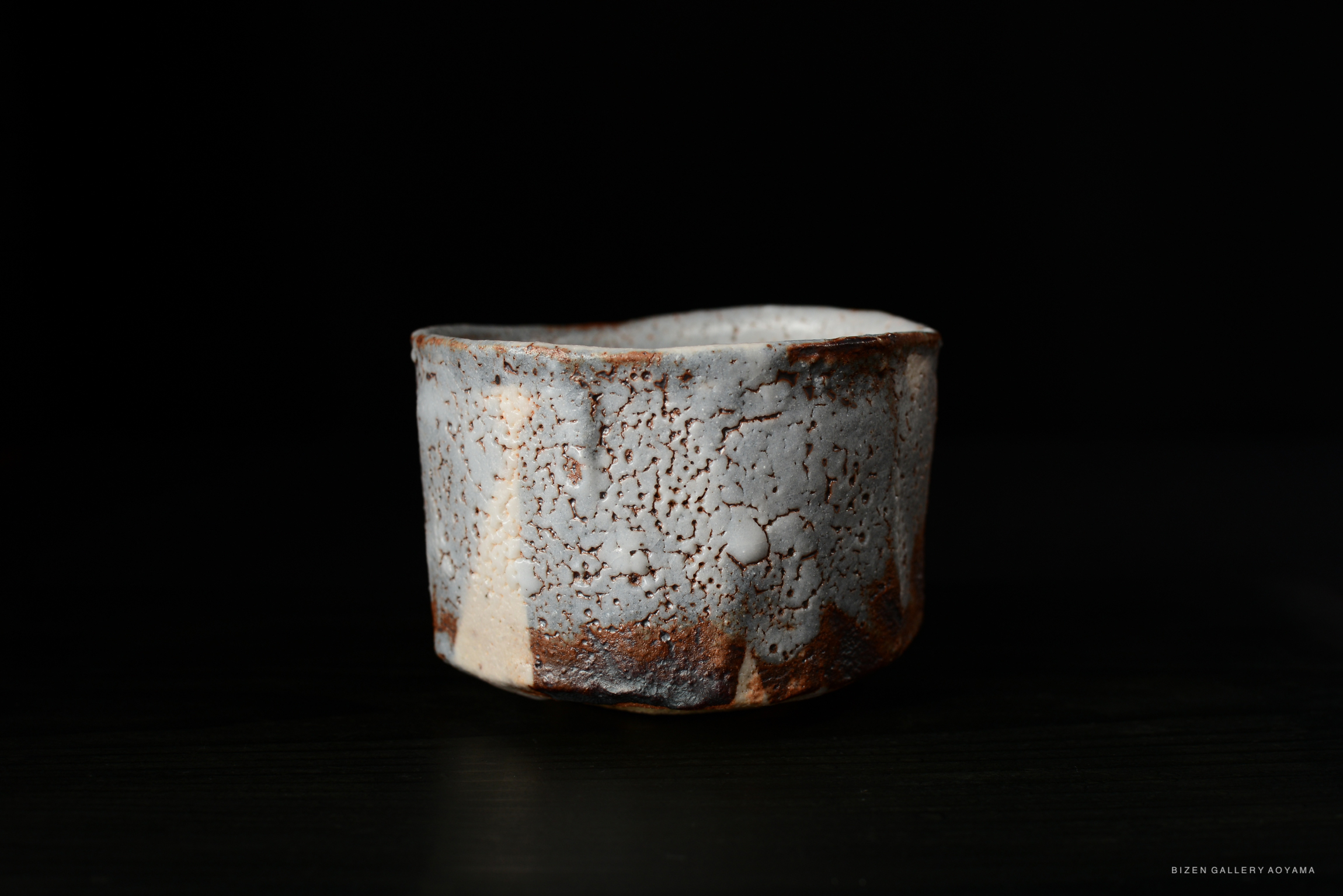 A side view of a Shino Chawan tea bowl, featuring a textured surface with cracks and a mix of light colors, set against a dark background.