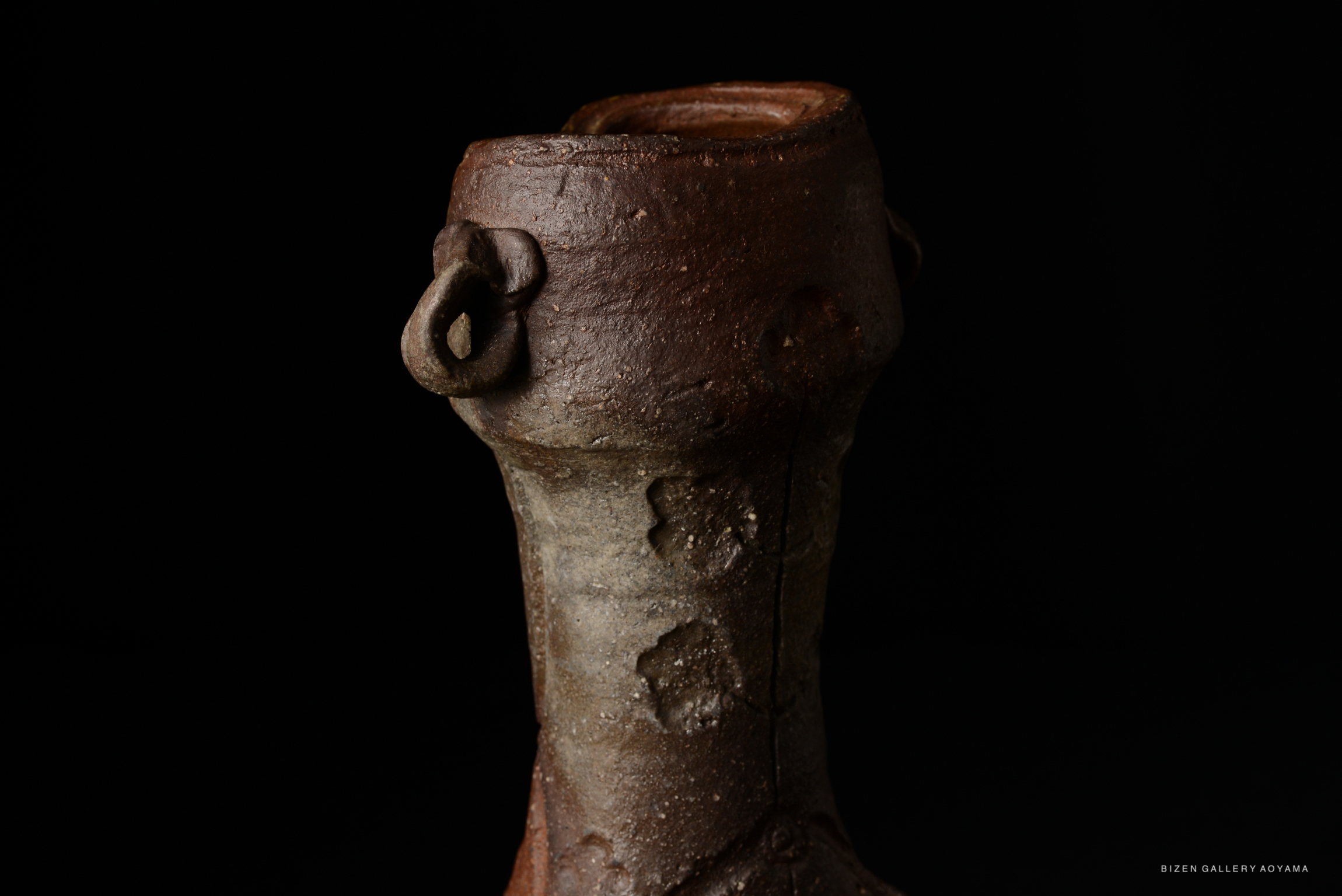 A close-up of a Bizen pottery vase with a distinctive dark, textured surface and unique handles, set against a black background.