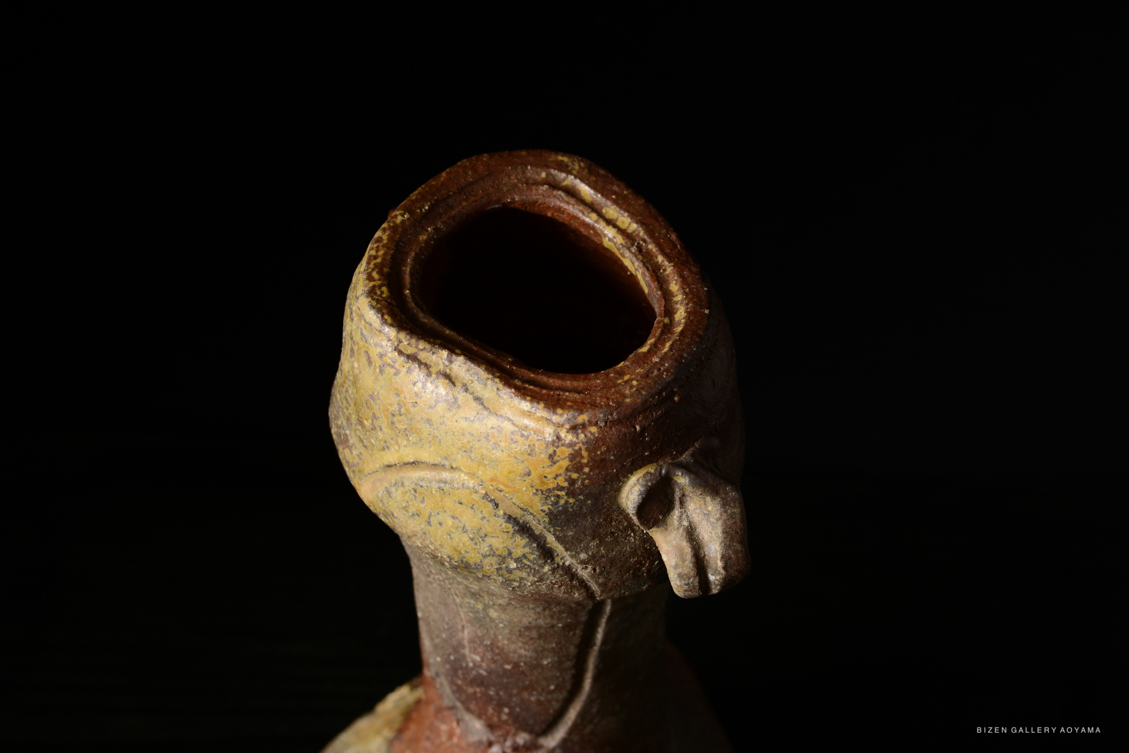 Close-up view of a Bizen pottery flower vase with a distinctive, textured surface and a narrow opening, set against a dark background.