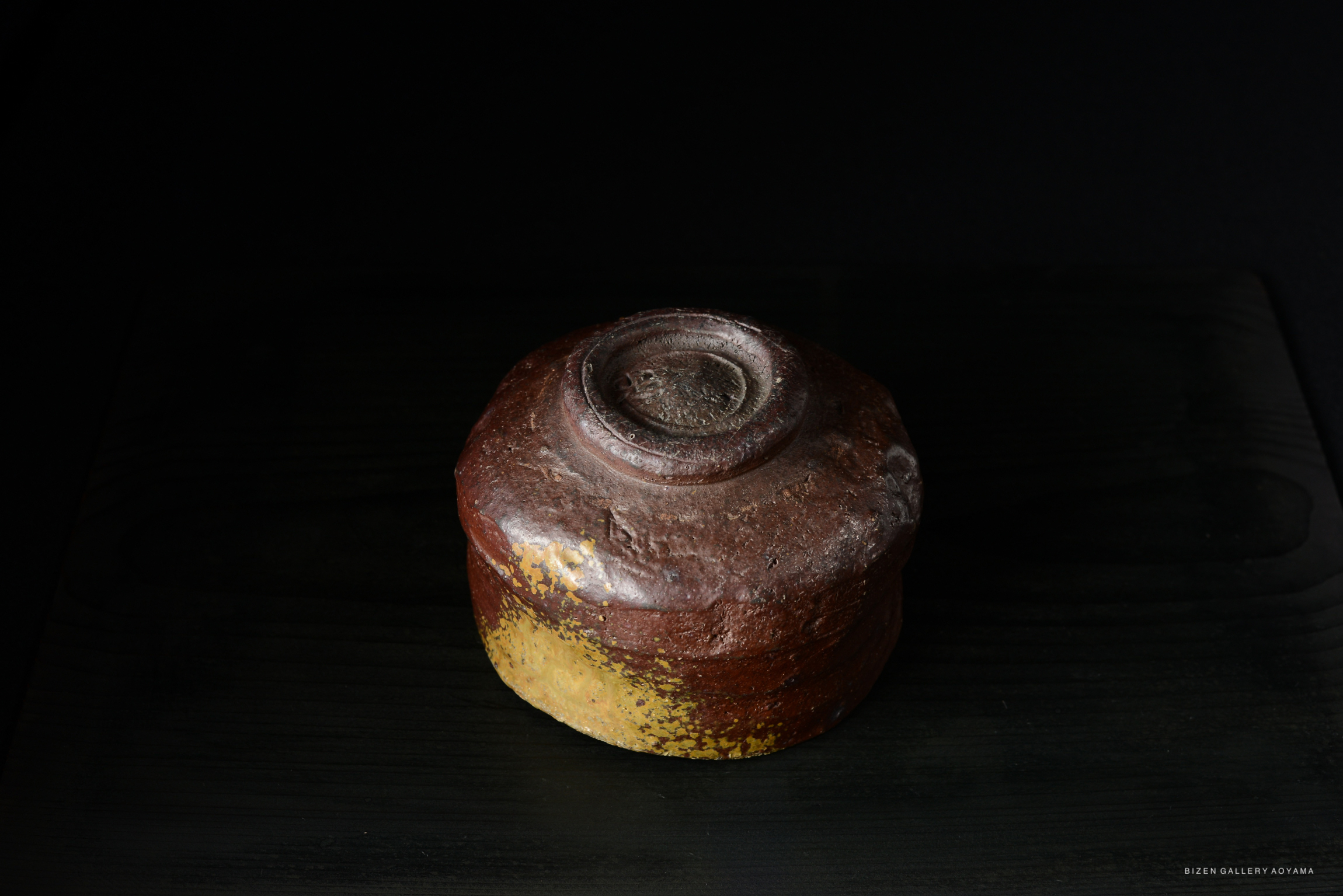 A ceramic bowl with a rough texture and a reddish-brown glaze, featuring a slightly yellowed base, set against a dark background.