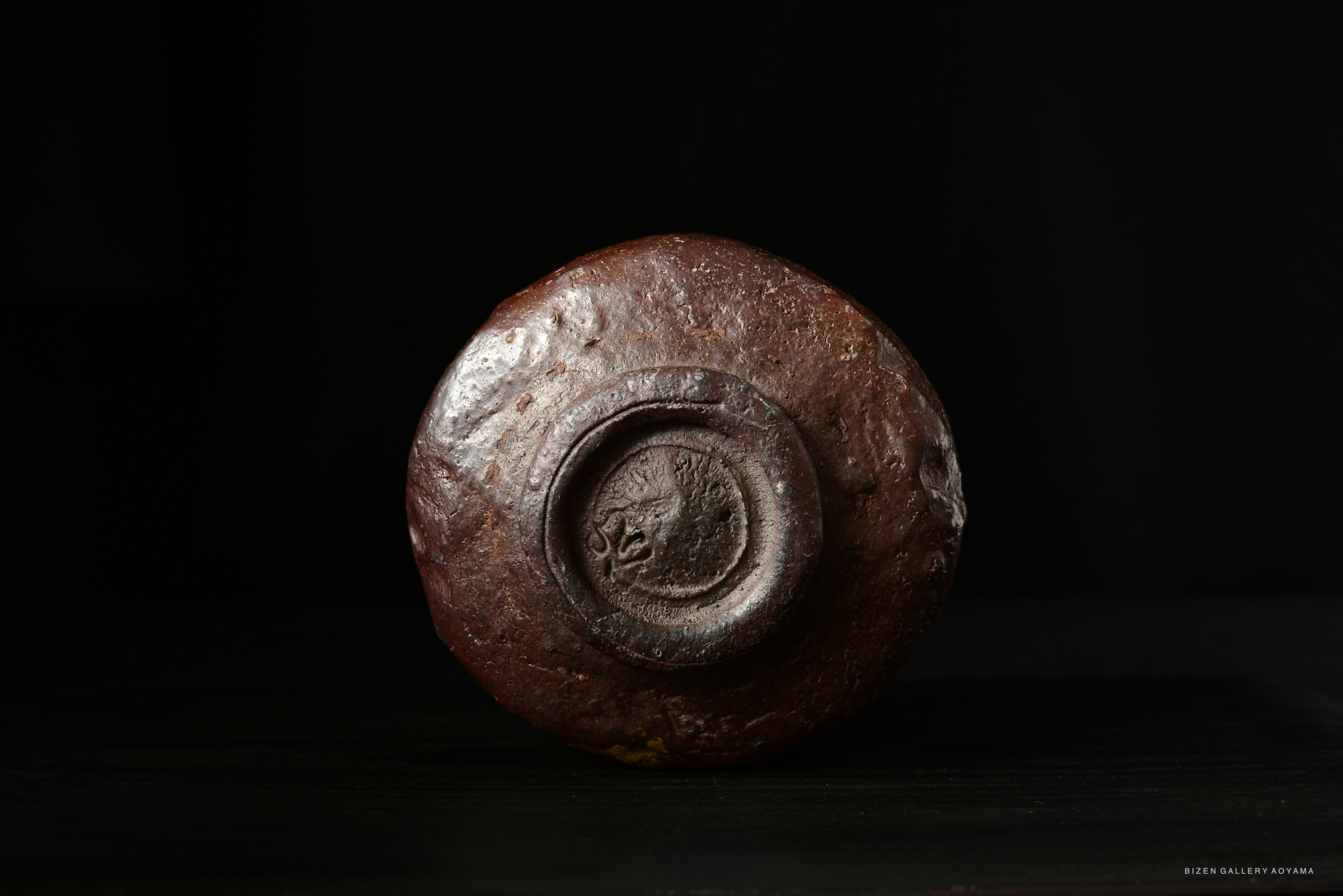 A close-up view of the bottom of a round, textured ceramic object with a small circular indentation, against a dark background.