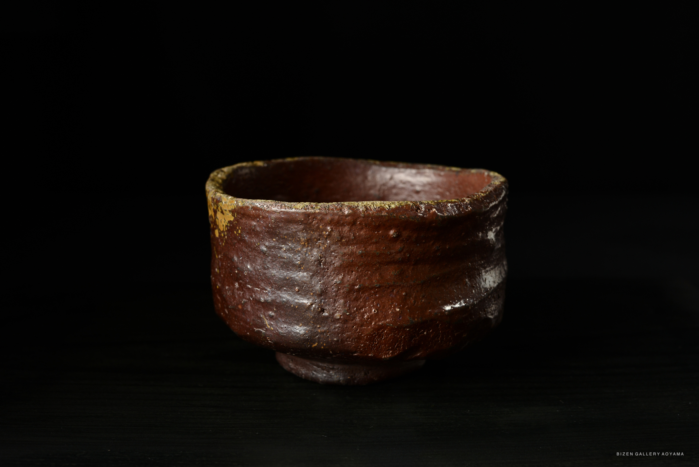 A rustic, handcrafted ceramic bowl with a rough, textured surface and a warm reddish-brown color, displayed against a dark background.