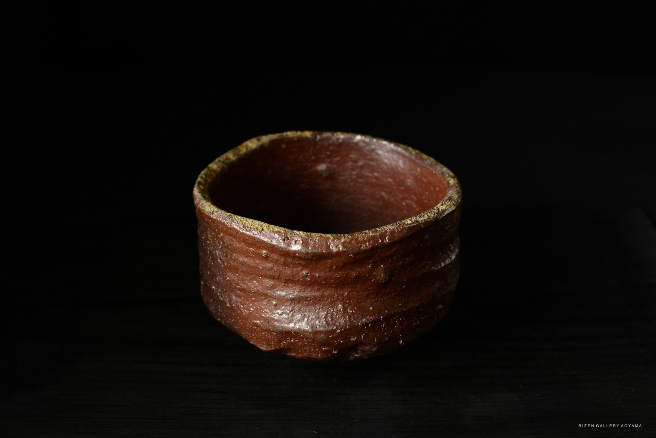 A handmade brown ceramic bowl with a slightly uneven rim, displayed against a dark background.