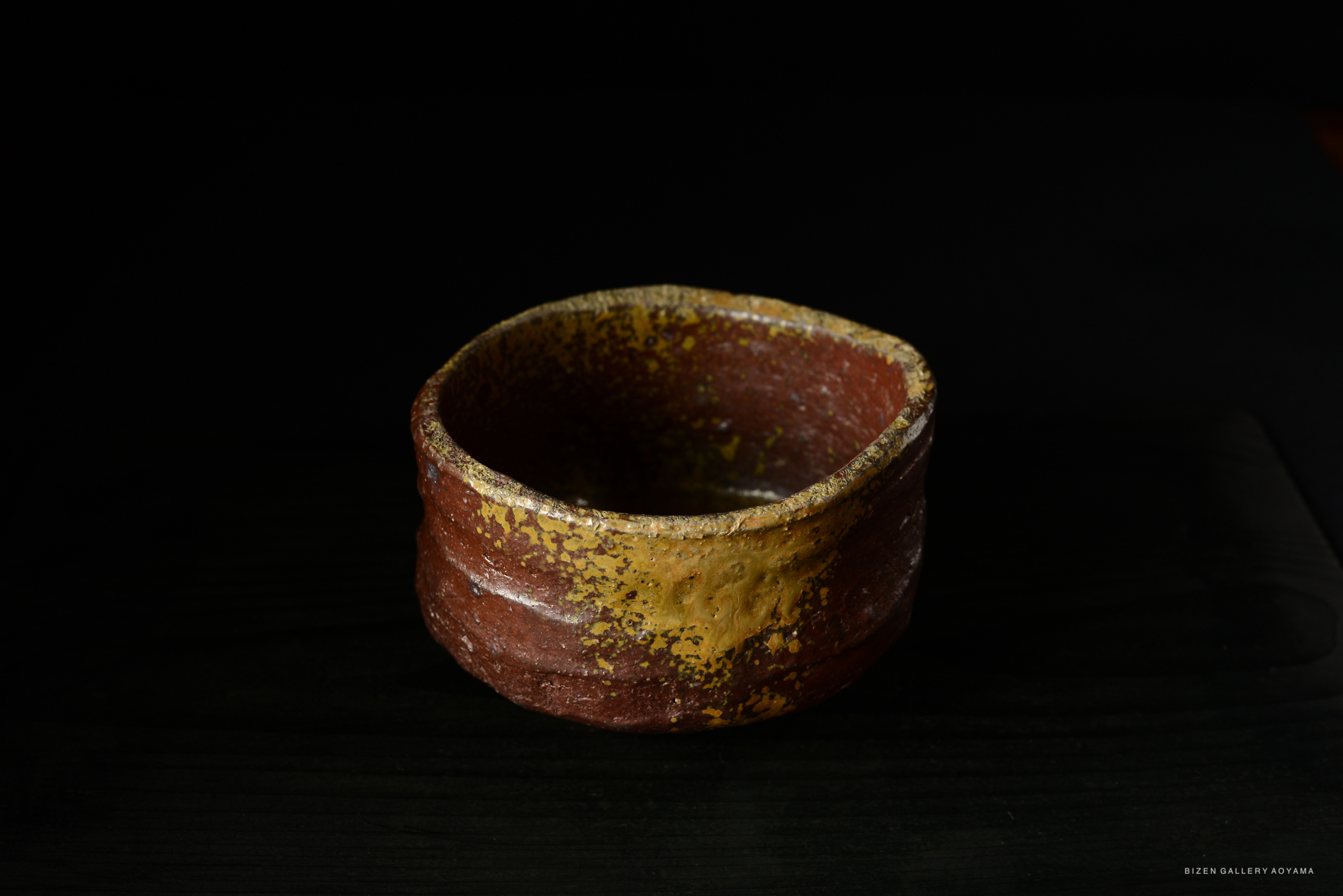 A close-up of a rustic, handmade pottery bowl with a reddish-brown exterior and a yellowish rim, placed on a dark surface.