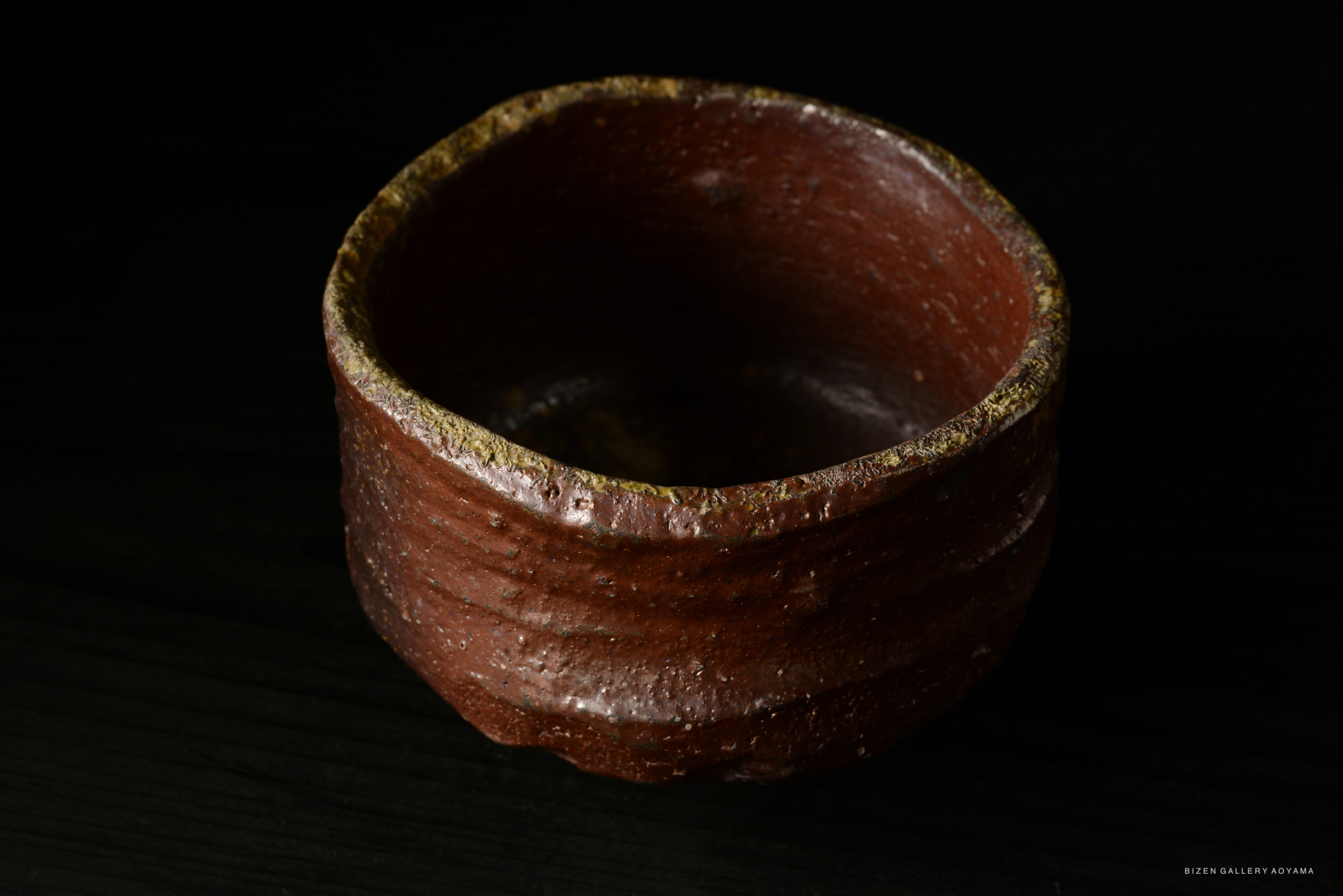 A close-up image of a rustic red clay bowl with a slightly textured surface and a wide opening, set against a dark background.