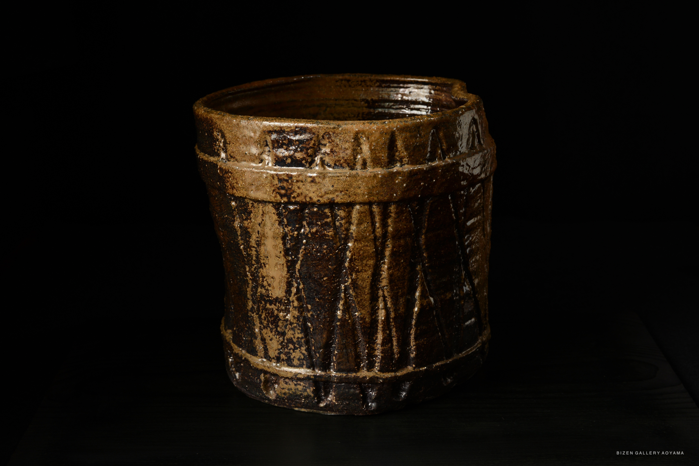 A traditional Bizen ware Mizusashi, showcasing a rustic design with textured patterns and earthy tones. The piece has a cylindrical shape and a slightly flared rim, set against a black background.