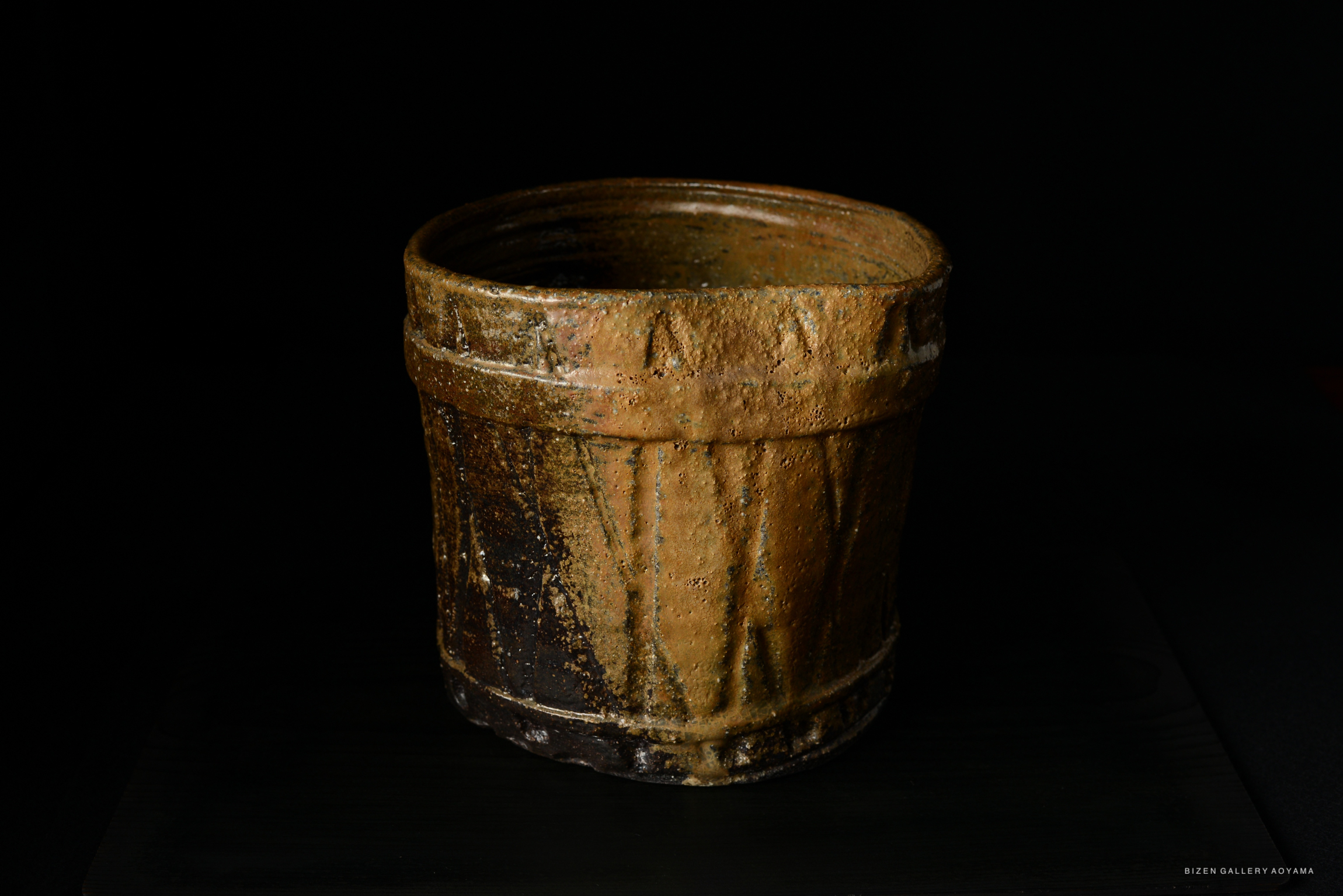 A rustic, round pottery vessel with a textured surface and earthy brown tones, set against a black background.