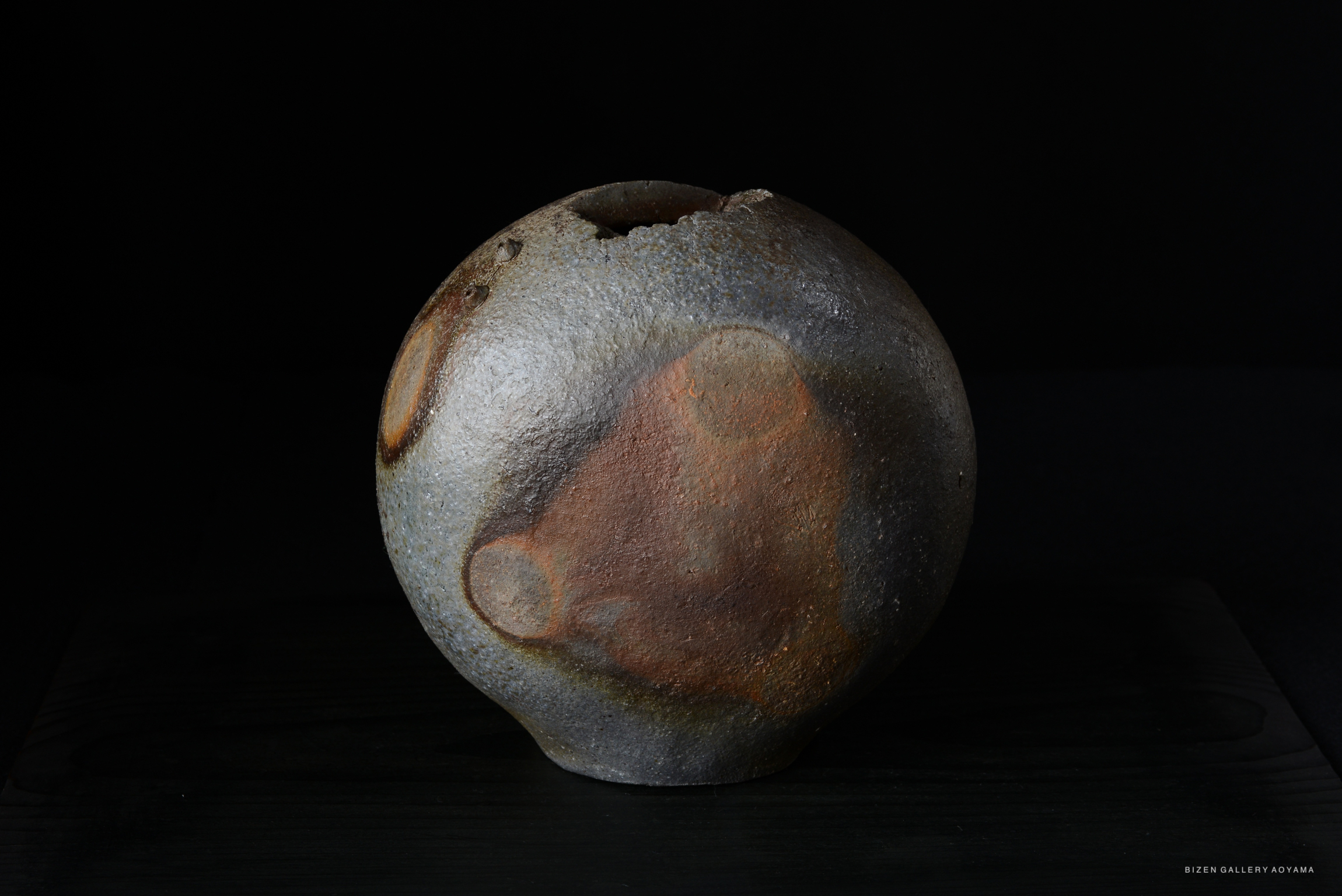 A round, rustic ceramic vase with a textured surface and earthy colors, displayed on a dark wooden base.