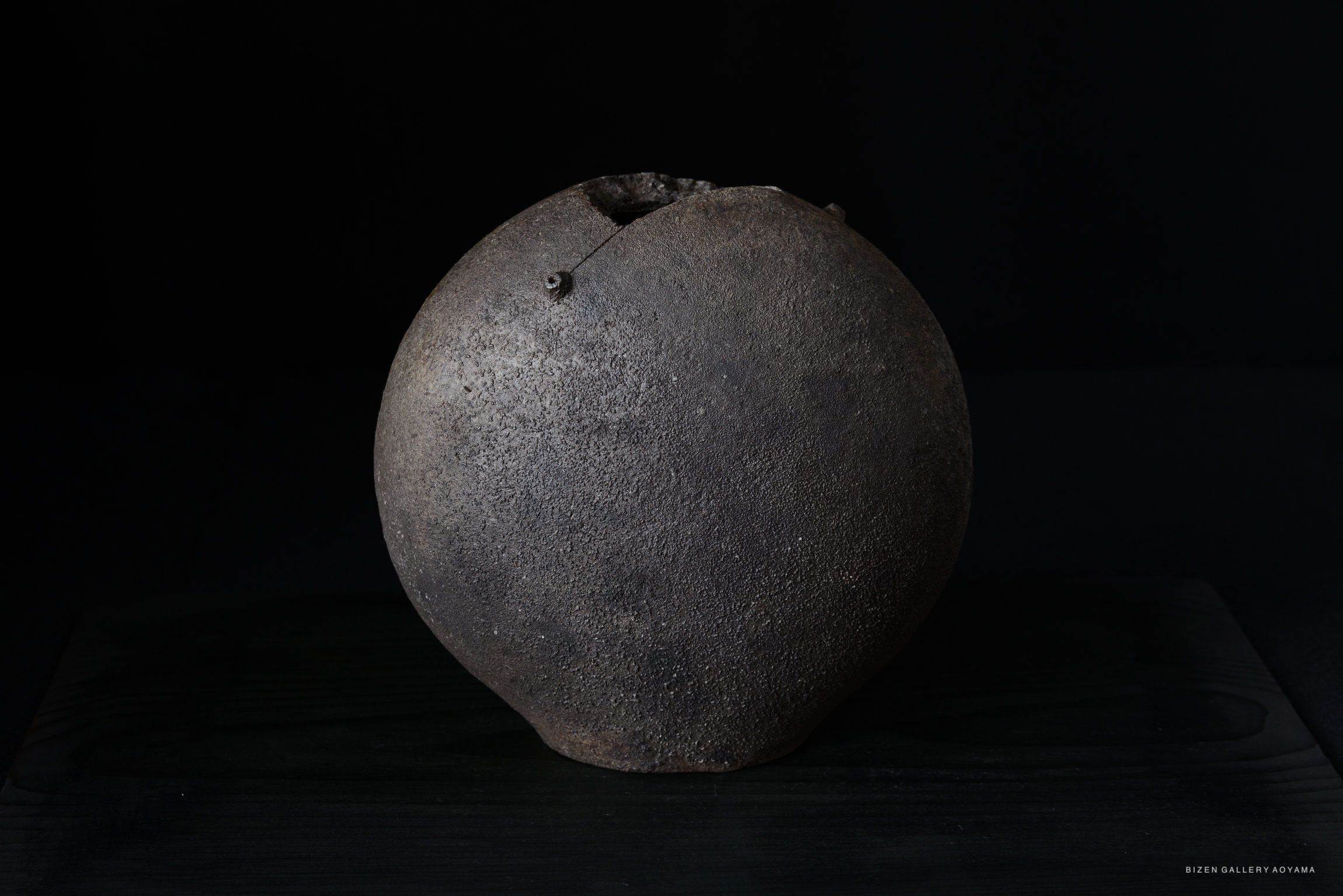 A round, textured ceramic vessel with a dark finish and a small opening at the top, displayed on a wooden surface against a black background.