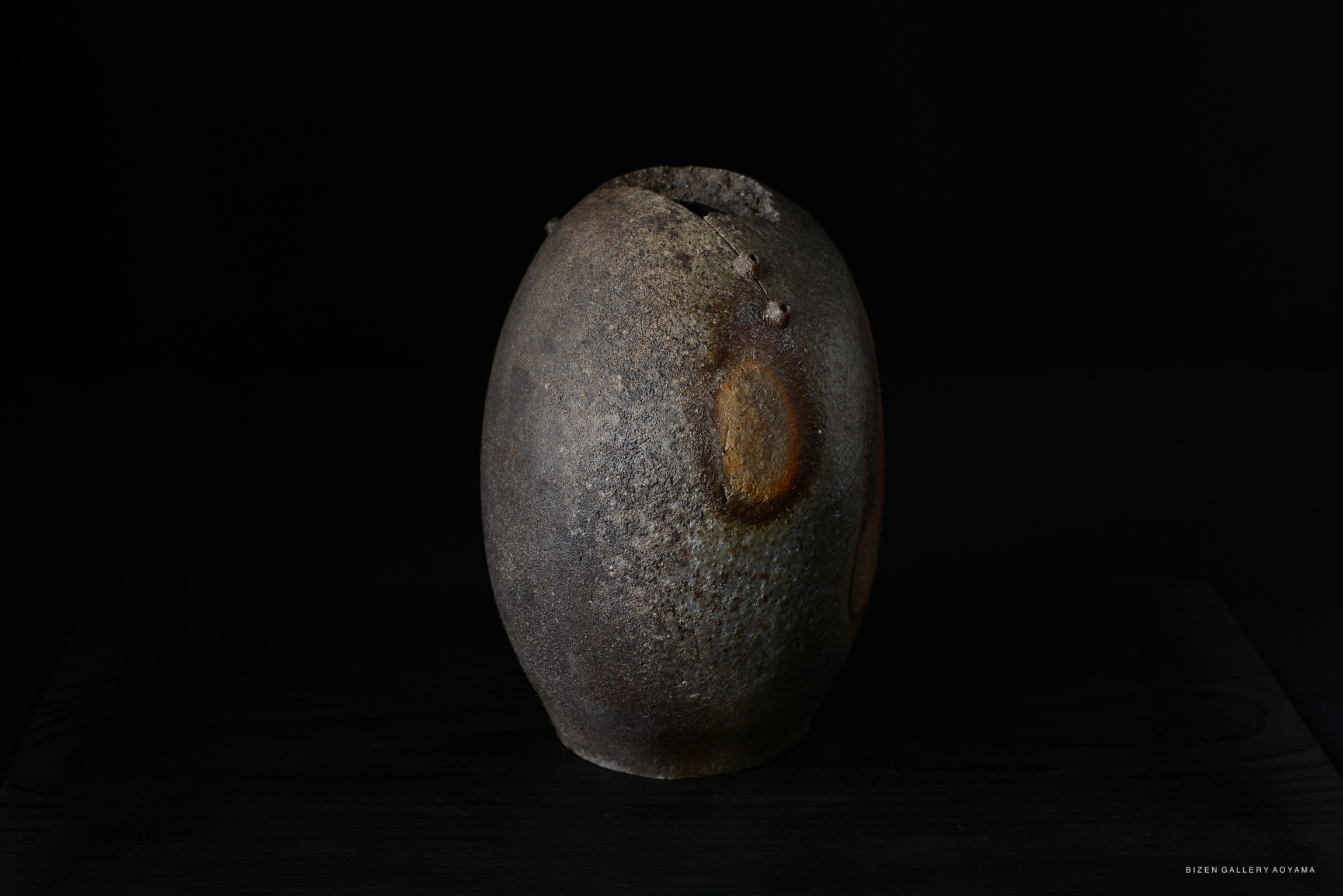 A rustic, round Bizen pottery vase with a textured surface and unique color variations, displayed against a dark background.