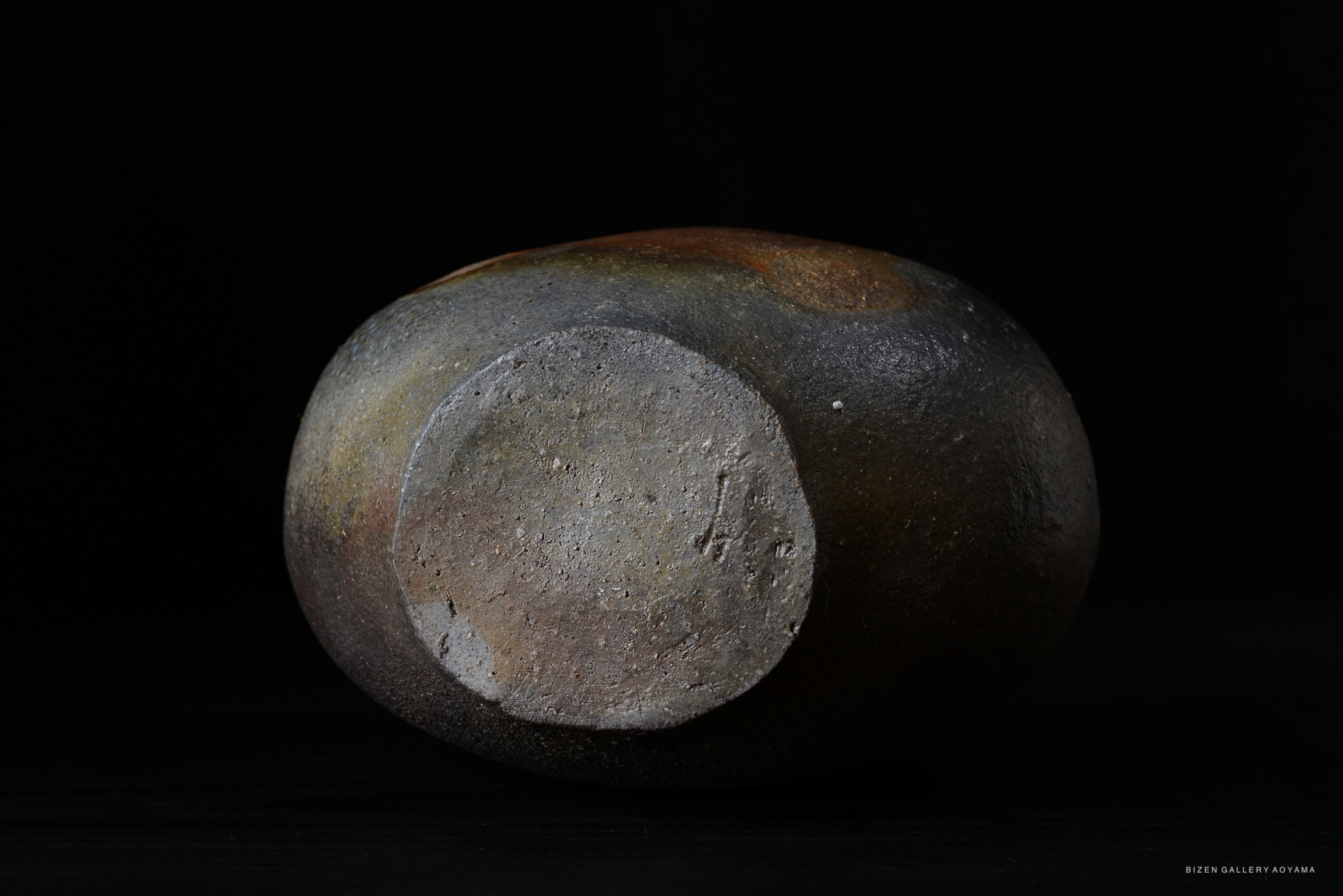 A round Bizen pottery vessel with a smooth, natural glaze and a flat base, displayed against a dark background.