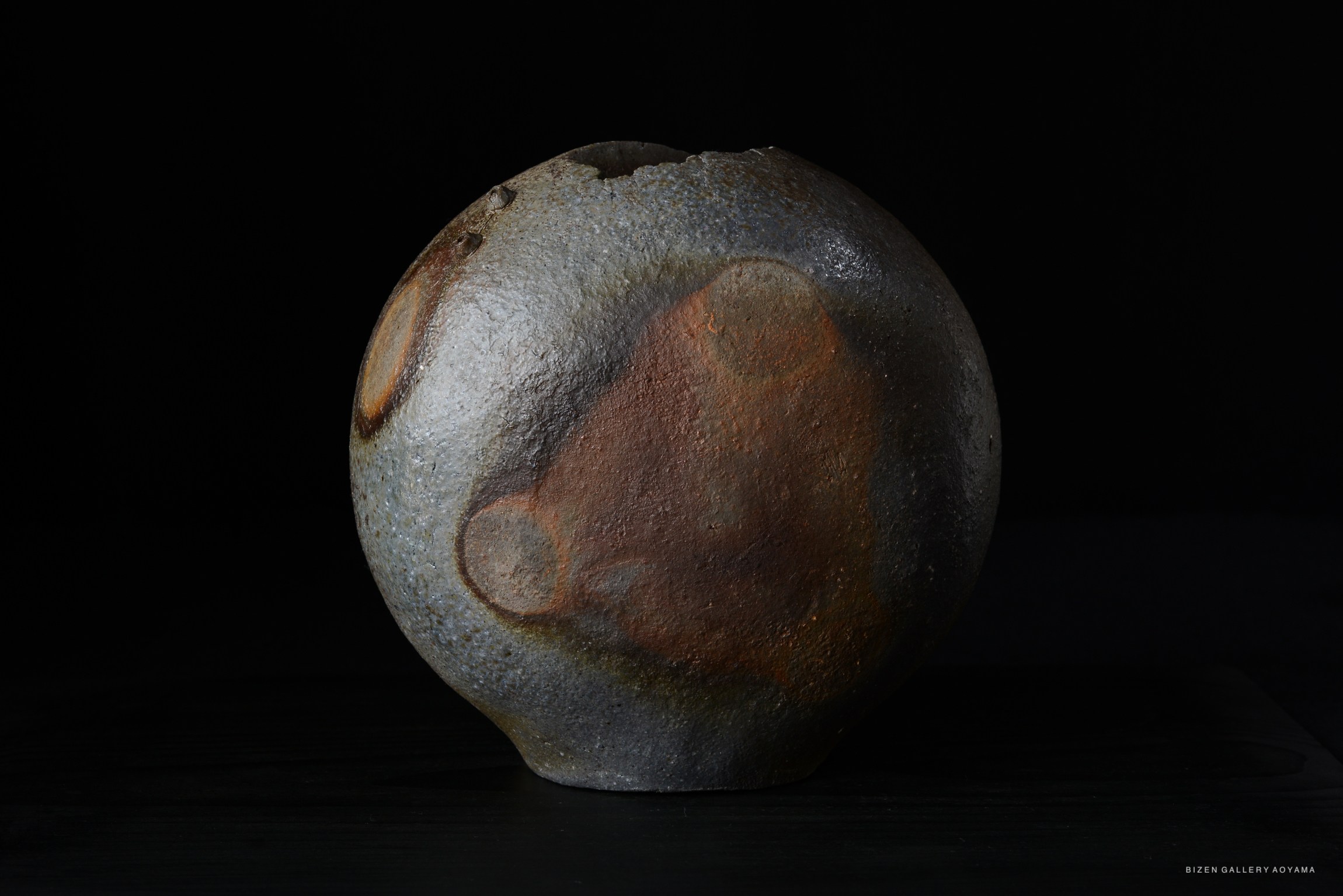 A unique Bizen ware ceramic piece with a spherical shape, showcasing a textured surface in deep gray and earthy colors, partially damaged at the top.