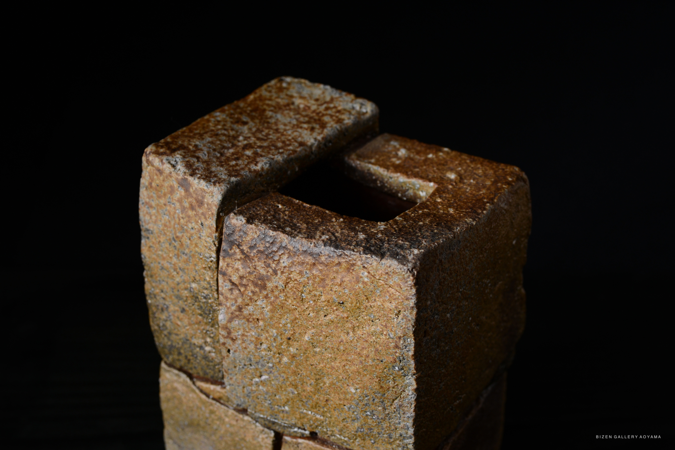 Close-up of a textured clay sculpture with a square opening on top, showcasing earthy tones against a dark background.