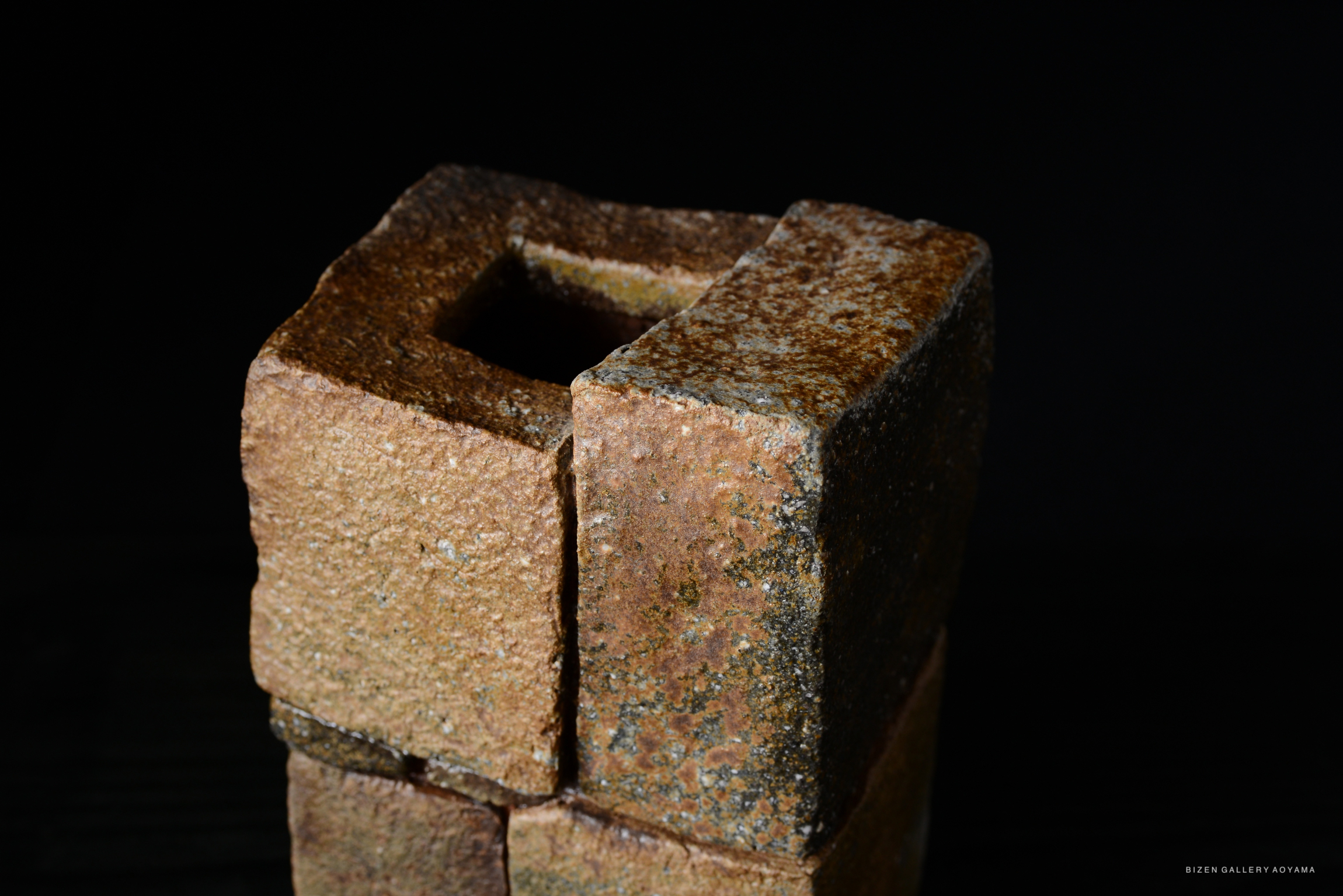 Close-up of a textured clay sculpture with a hollow square opening on top, set against a dark background.