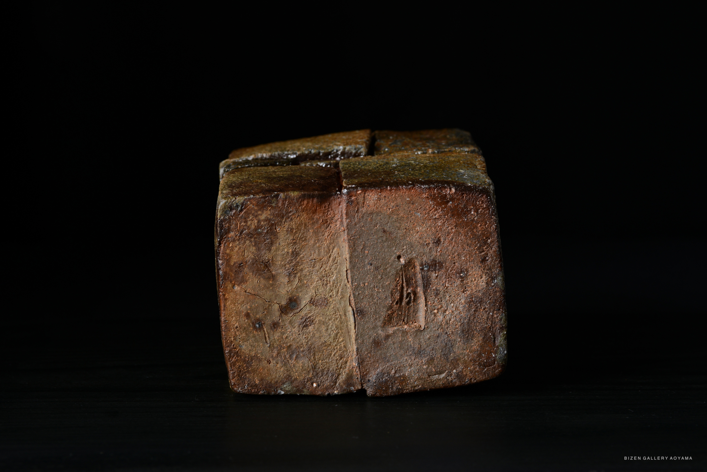 A rustic, square stoneware piece with a textured, earthy surface, showcasing a natural glaze and subtle markings, set against a dark background.