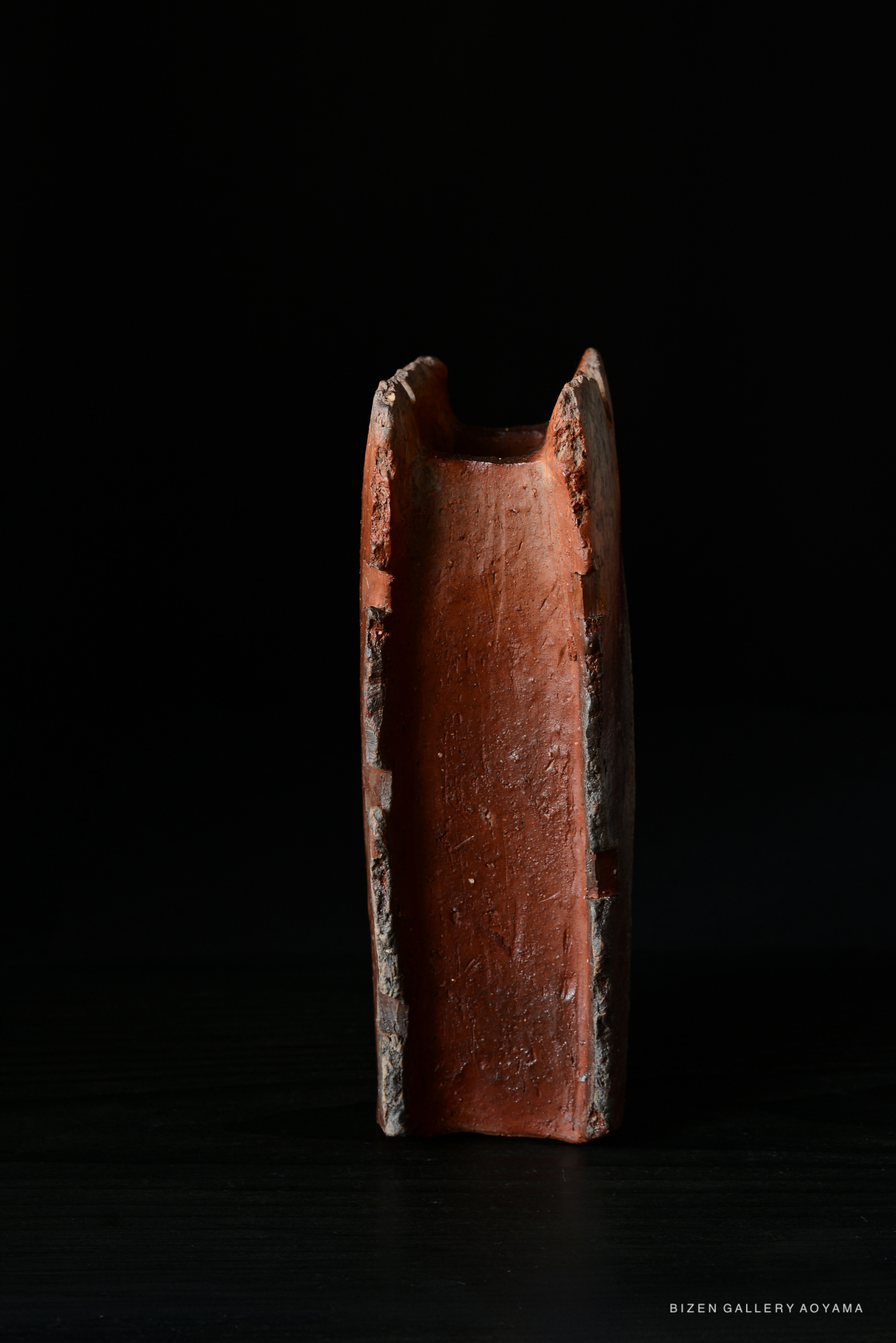 A vertical piece of Bizen pottery with a smooth reddish surface and rough edges, set against a black background.