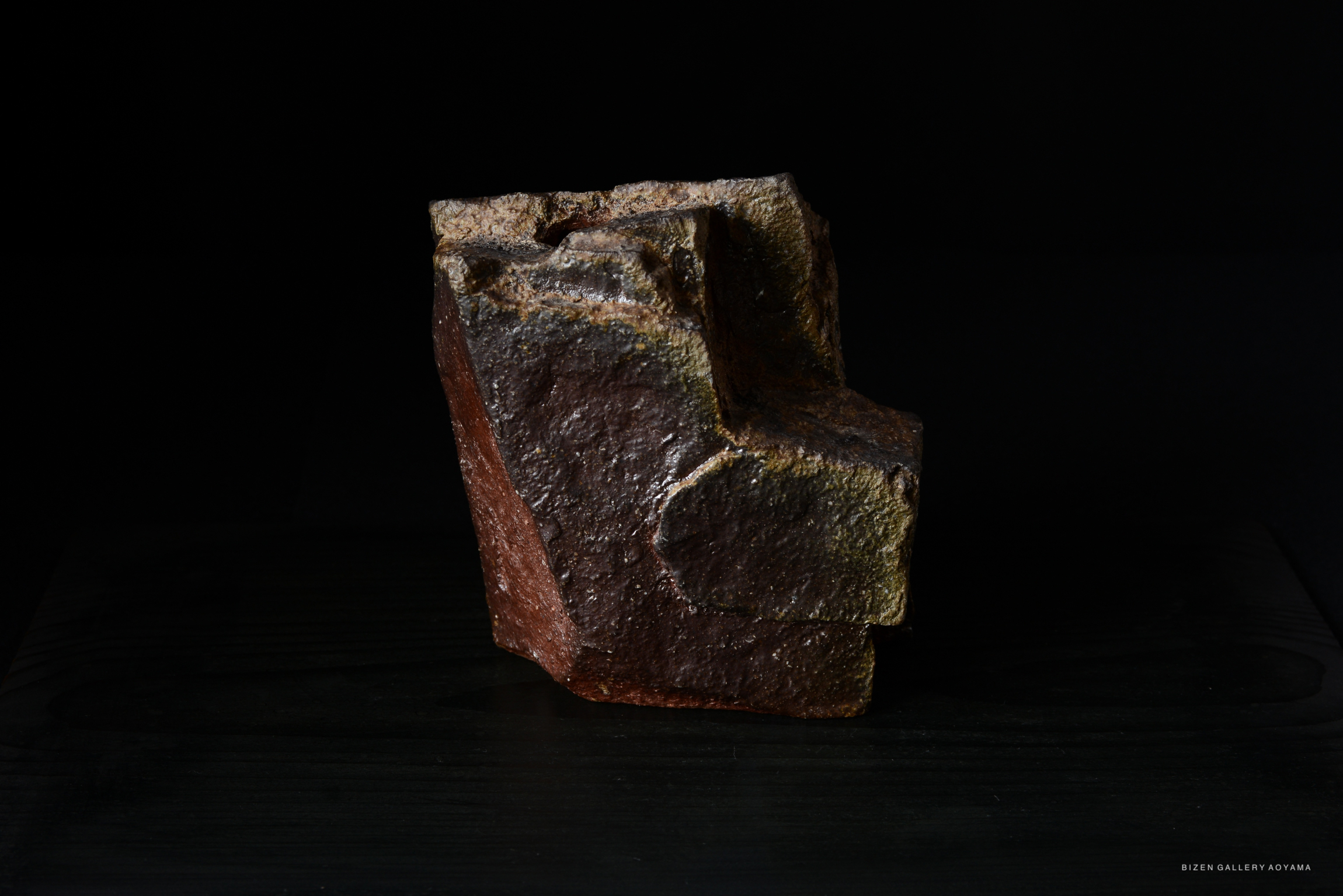 A textured ceramic sculpture with earthy tones, displayed on a dark surface against a black background.