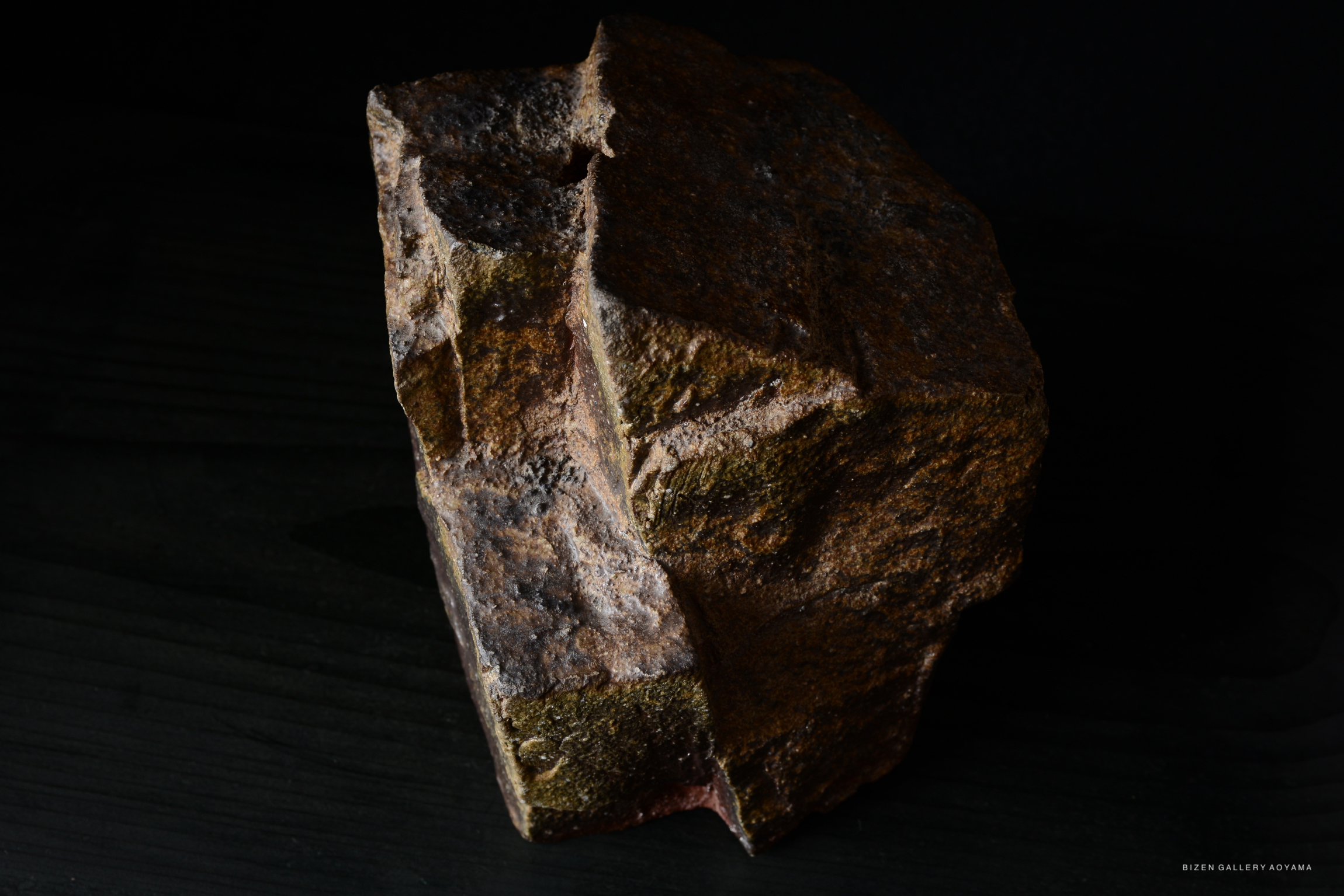 A textured, irregularly shaped rock with earthy tones of brown and hints of green, set against a dark background.