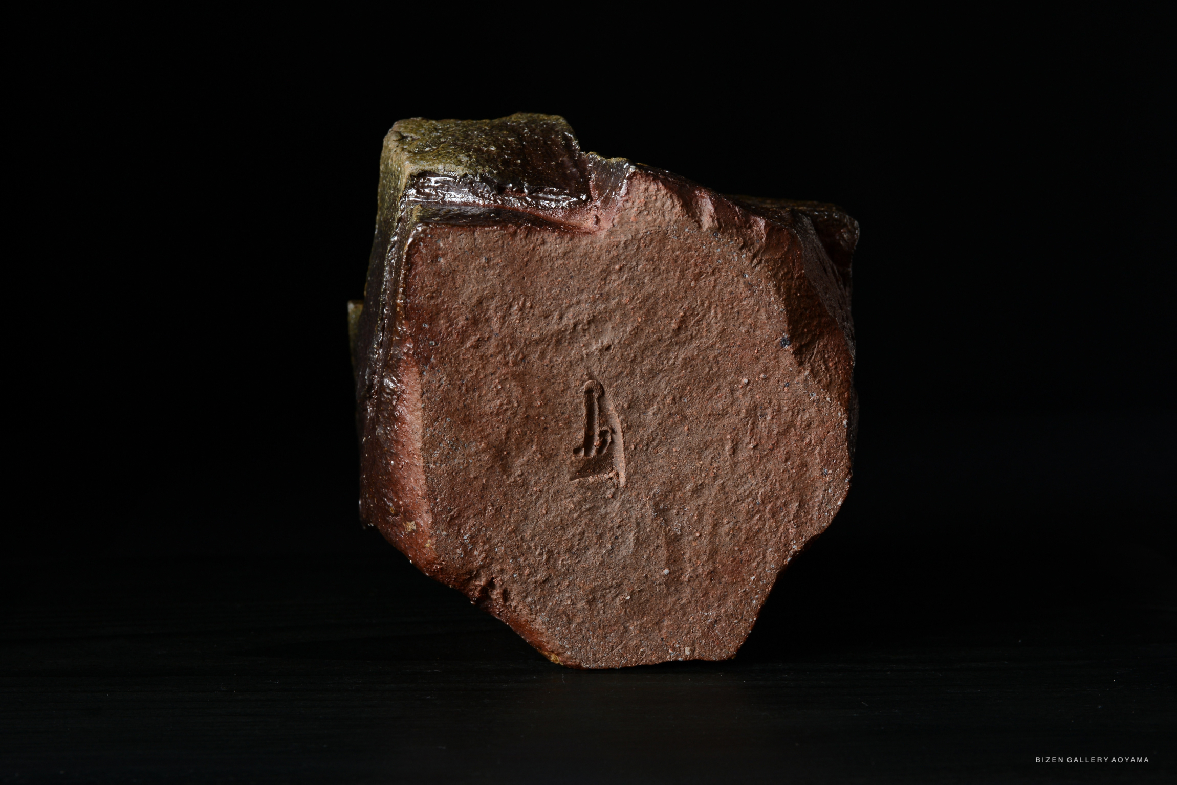 A close-up view of a textured, dark ceramic object with a visible carving on its surface, set against a black background.