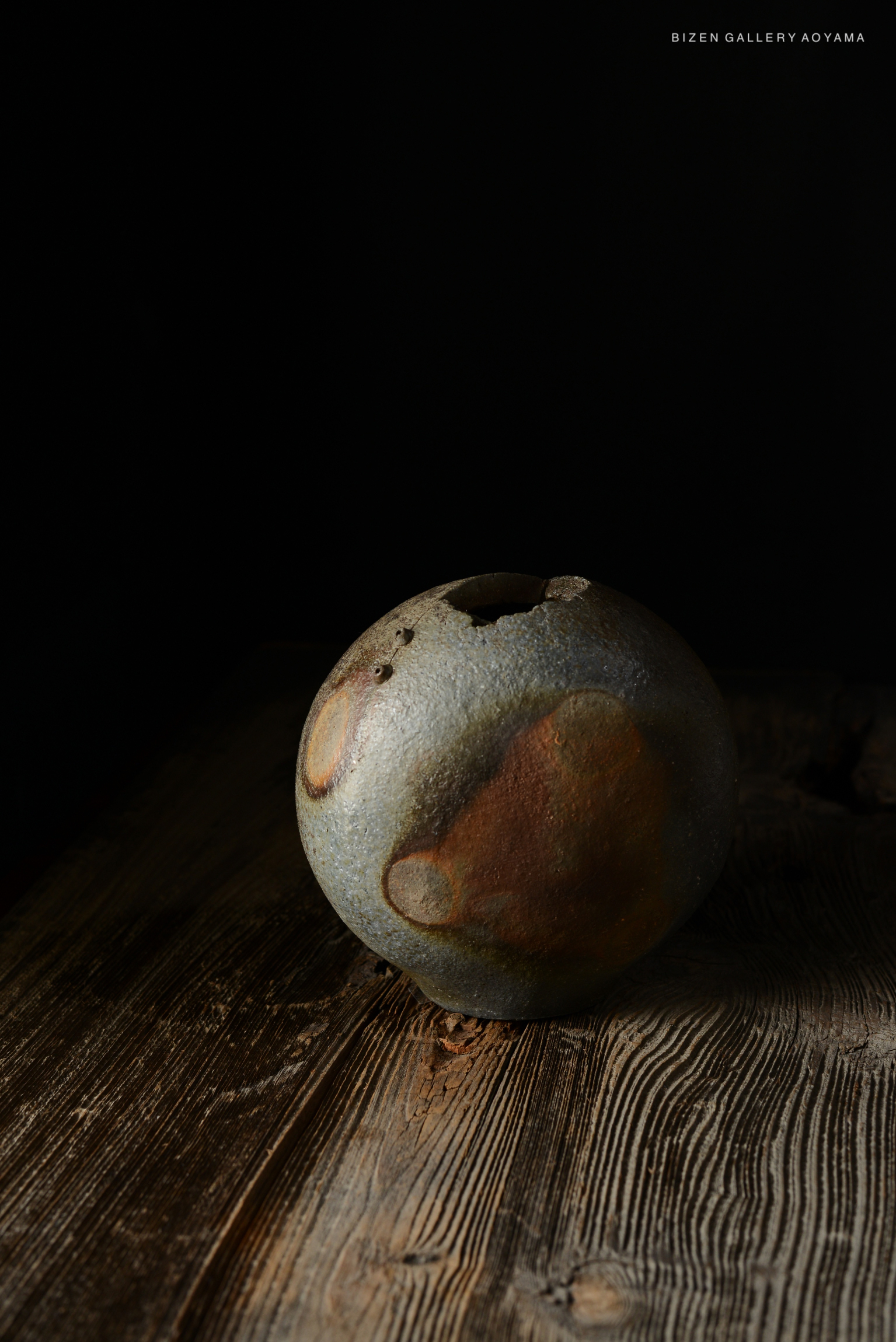 Round clay vase with a textured surface and earthy tones, placed on a wooden table against a dark background.