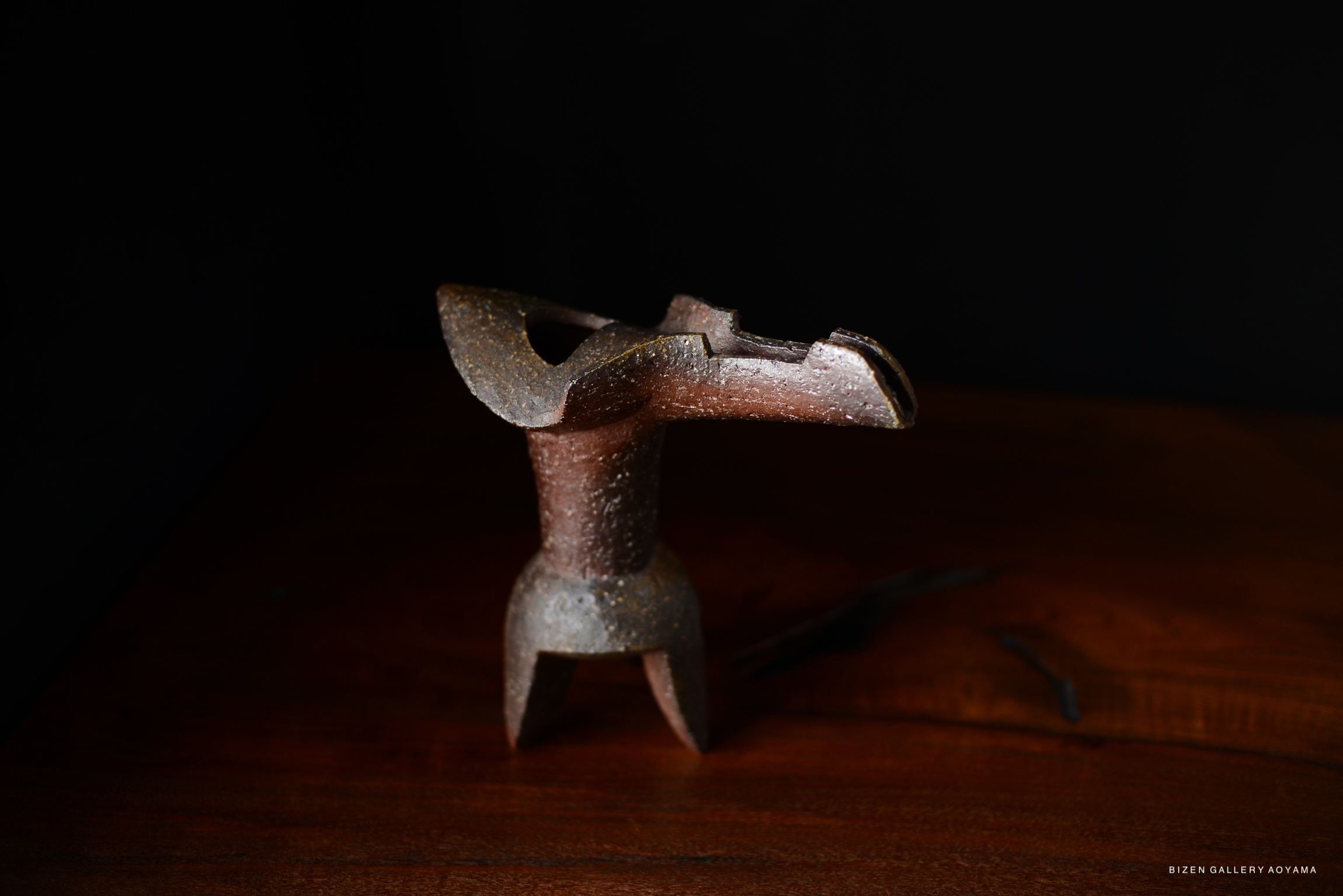 A unique Hanaire Shaku ceramic vase with a textured brown surface, designed for flower arrangement, displayed on a wooden surface against a dark background.