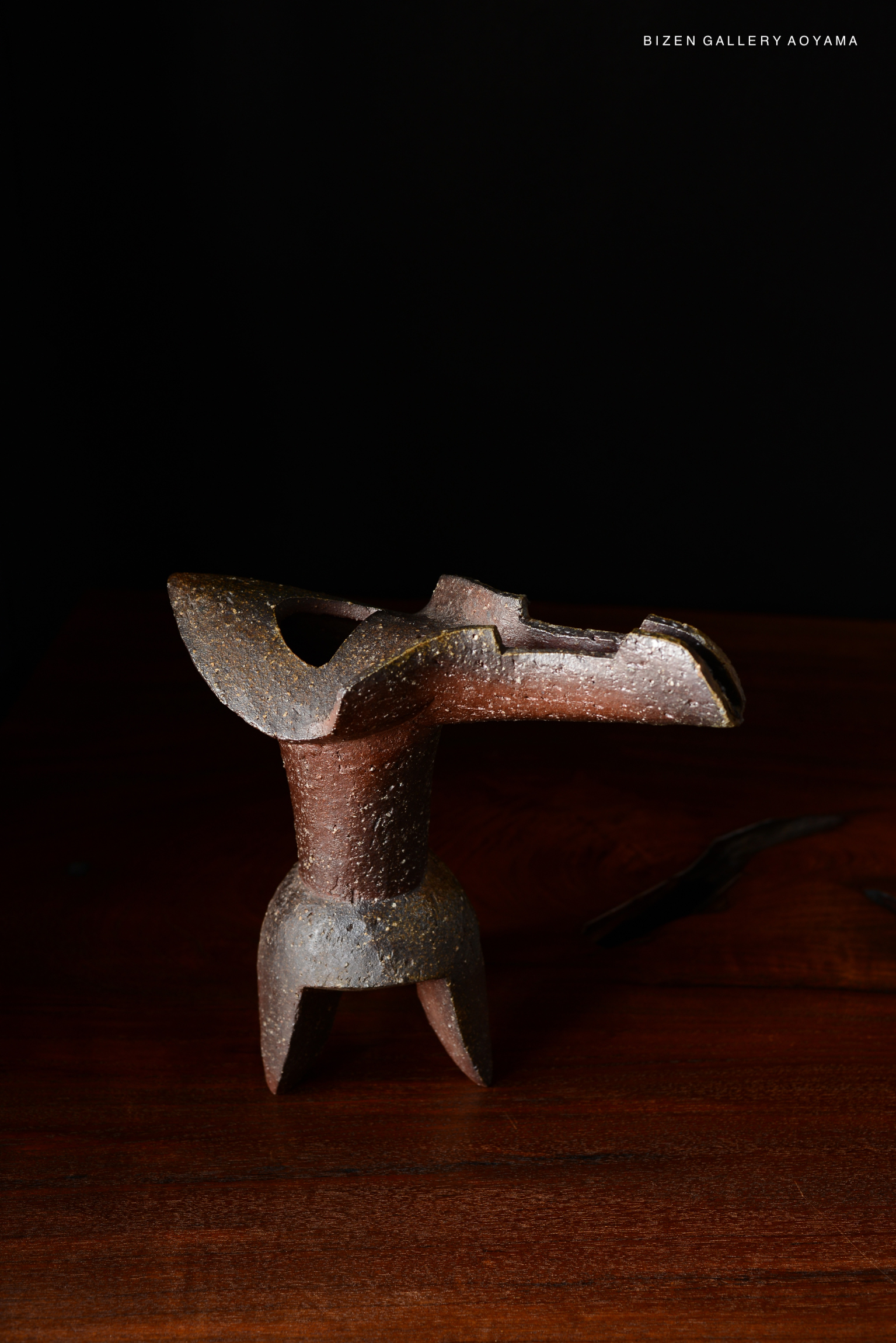 A unique hanaire vase made of ceramic, featuring a textured, brownish surface and a distinctive shape, displayed on a wooden table against a dark background.