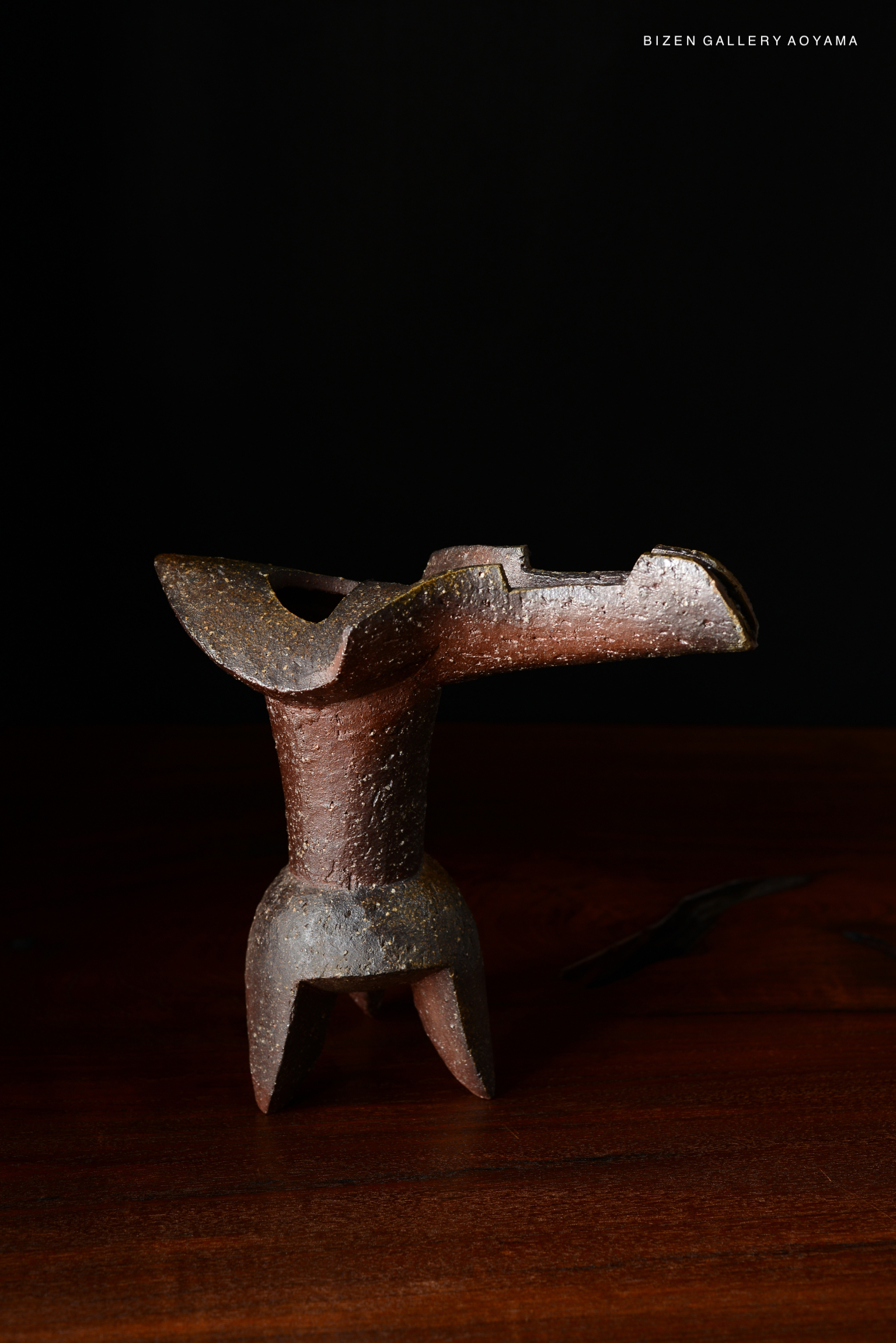 A unique ceramic hanaire (flower vase) with a rustic design, featuring a reddish-brown hue and textured surface, displayed on a wooden table against a dark background.