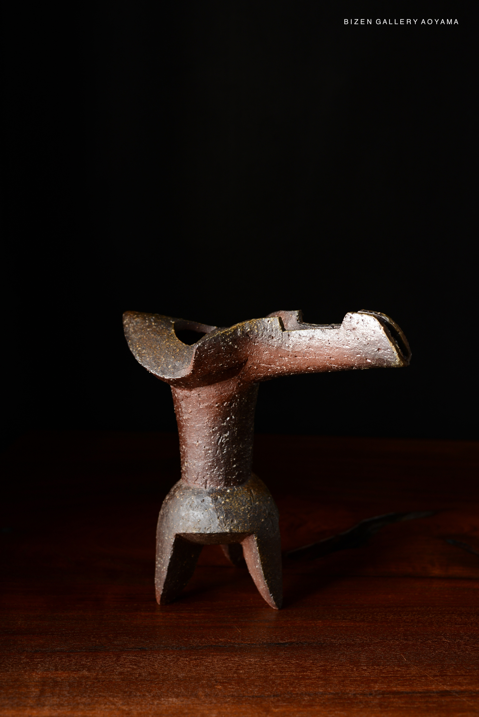 A rustic Bizen pottery vase, featuring a unique design with a curved neck and a textured surface, displayed on a dark wooden table against a black background.