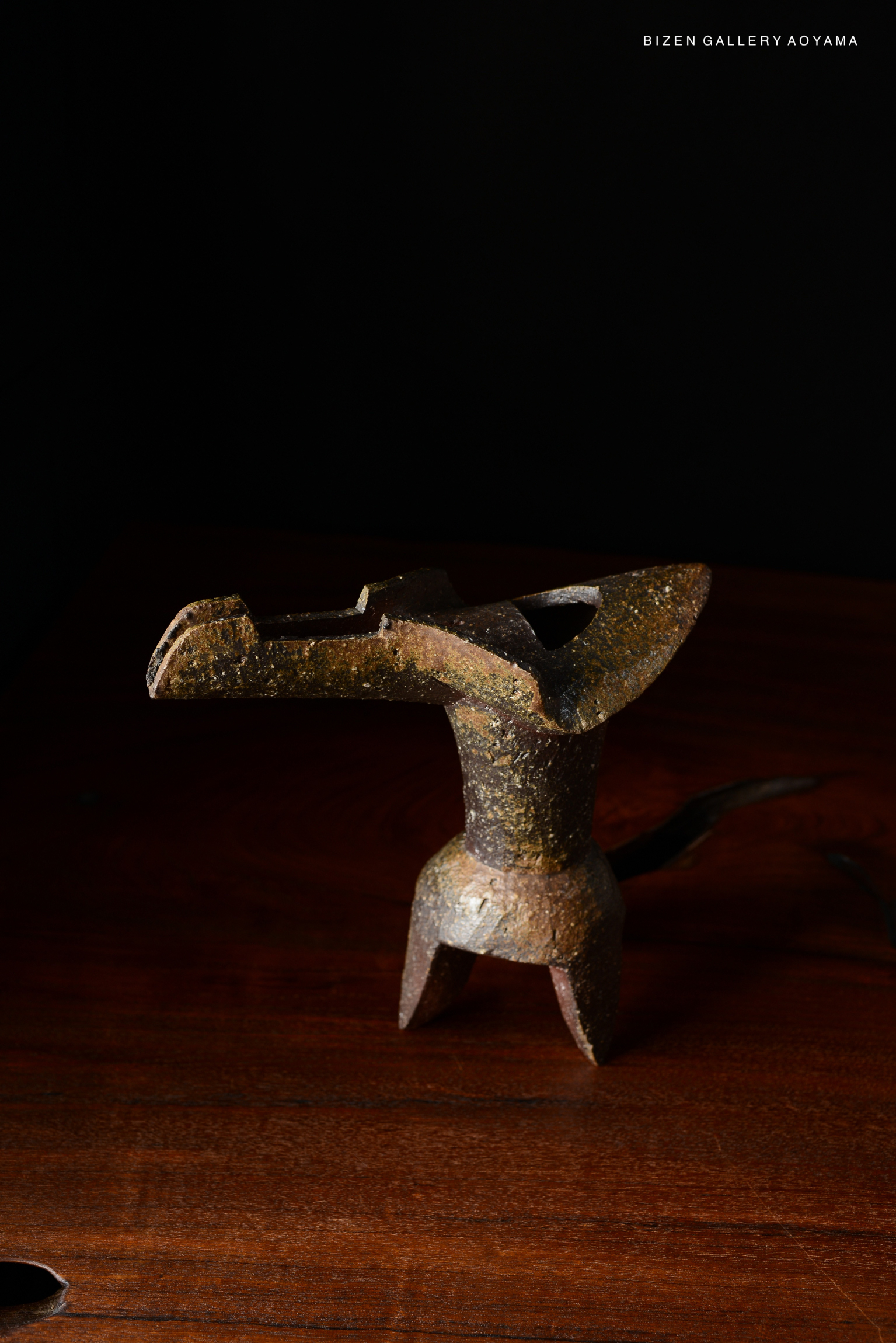 A decorative metal vase holder with a unique design, set against a dark background. The object has a rustic finish and stands upright on three legs.