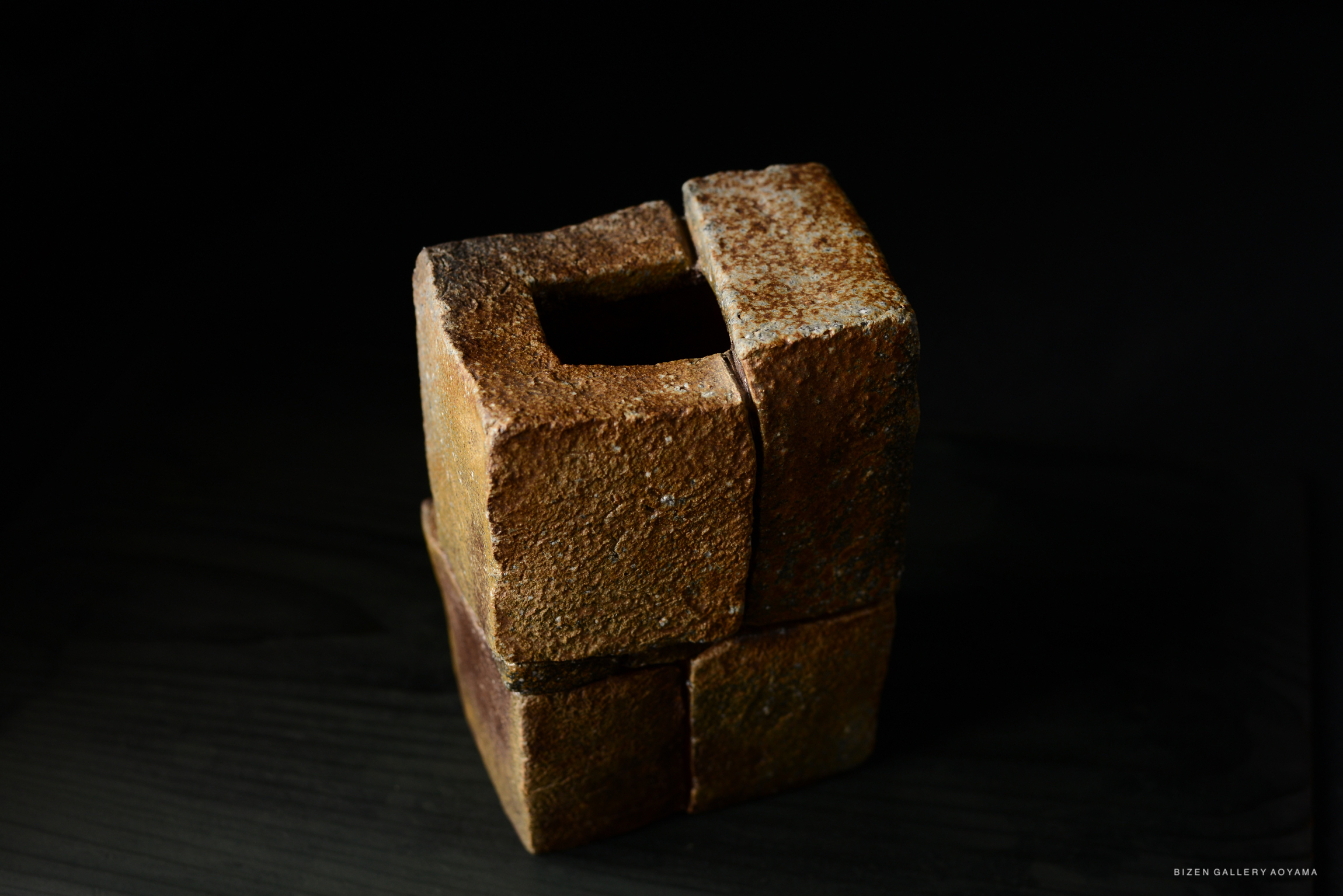 A close-up view of a unique ceramic flower vase with a square, stacked design, featuring earthy tones and a textured surface against a dark background.