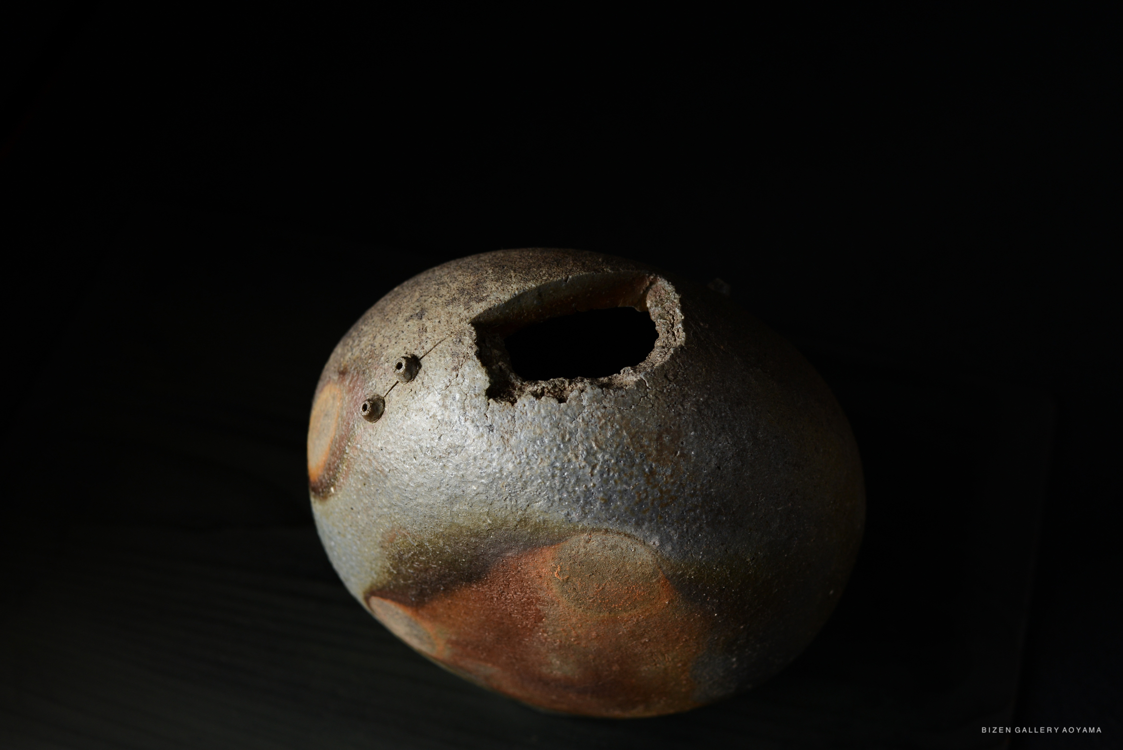 A unique Bizen pottery vase with a round shape, featuring a rough texture and earthy tones, set against a dark background.