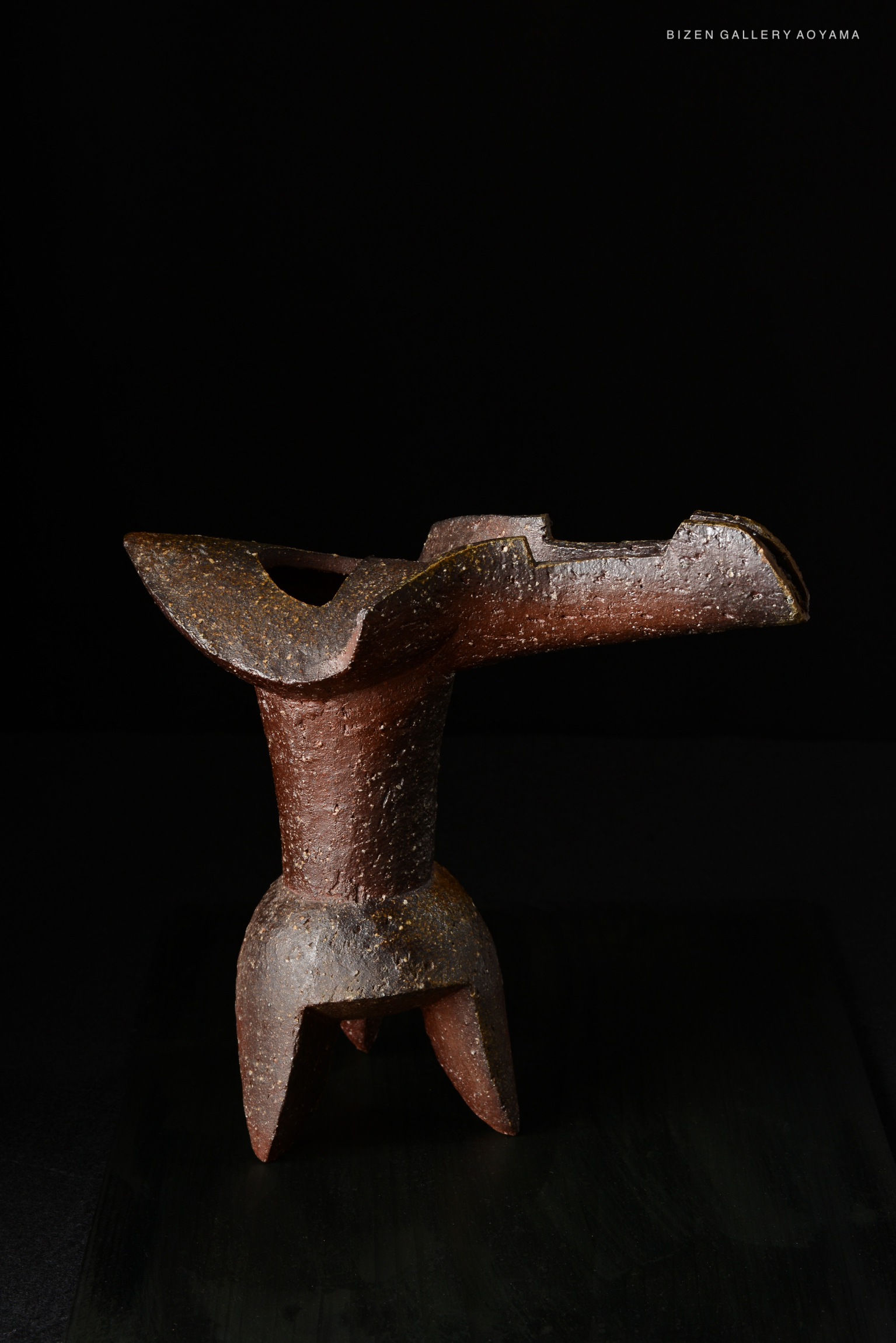 A unique ceramic vase with a sculptural design, featuring earthy tones and a textured surface, displayed against a dark background.