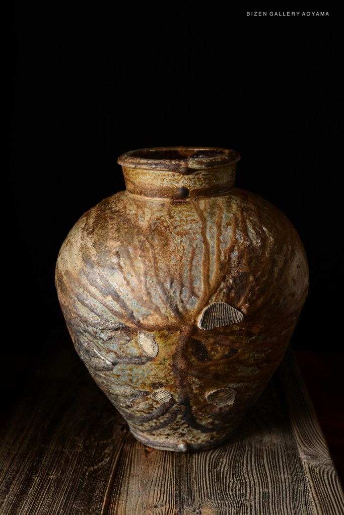 A close-up view of a Bizen pottery vase with a textured surface and natural glaze, displayed on a wooden table against a dark background.