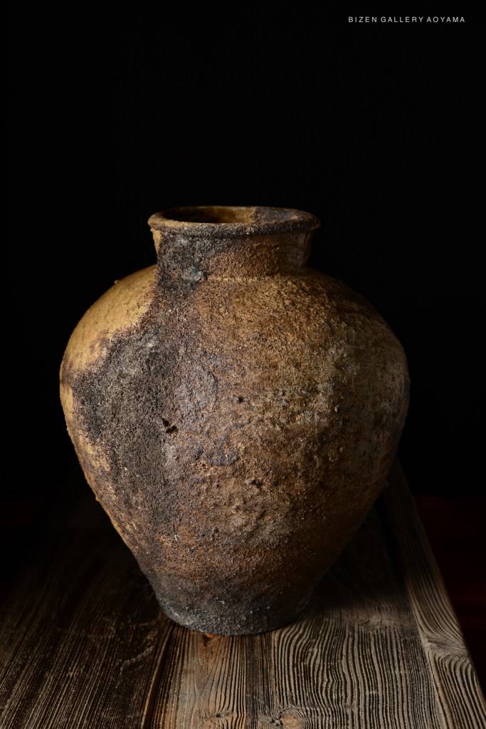 A close-up view of a traditional Bizen pottery vase featuring a textured surface in earthy tones, displayed on a wooden table against a black background.