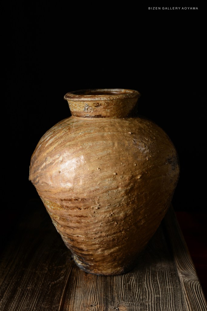 Close-up of a Bizen pottery vase with a textured surface and natural earth tones, placed on a wooden surface against a black background.