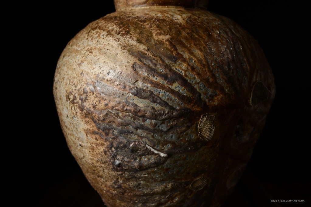 Close-up view of a Bizen pottery vase, showcasing its textured surface with intricate patterns and a rich, earthy color. The vase is set against a dark background.