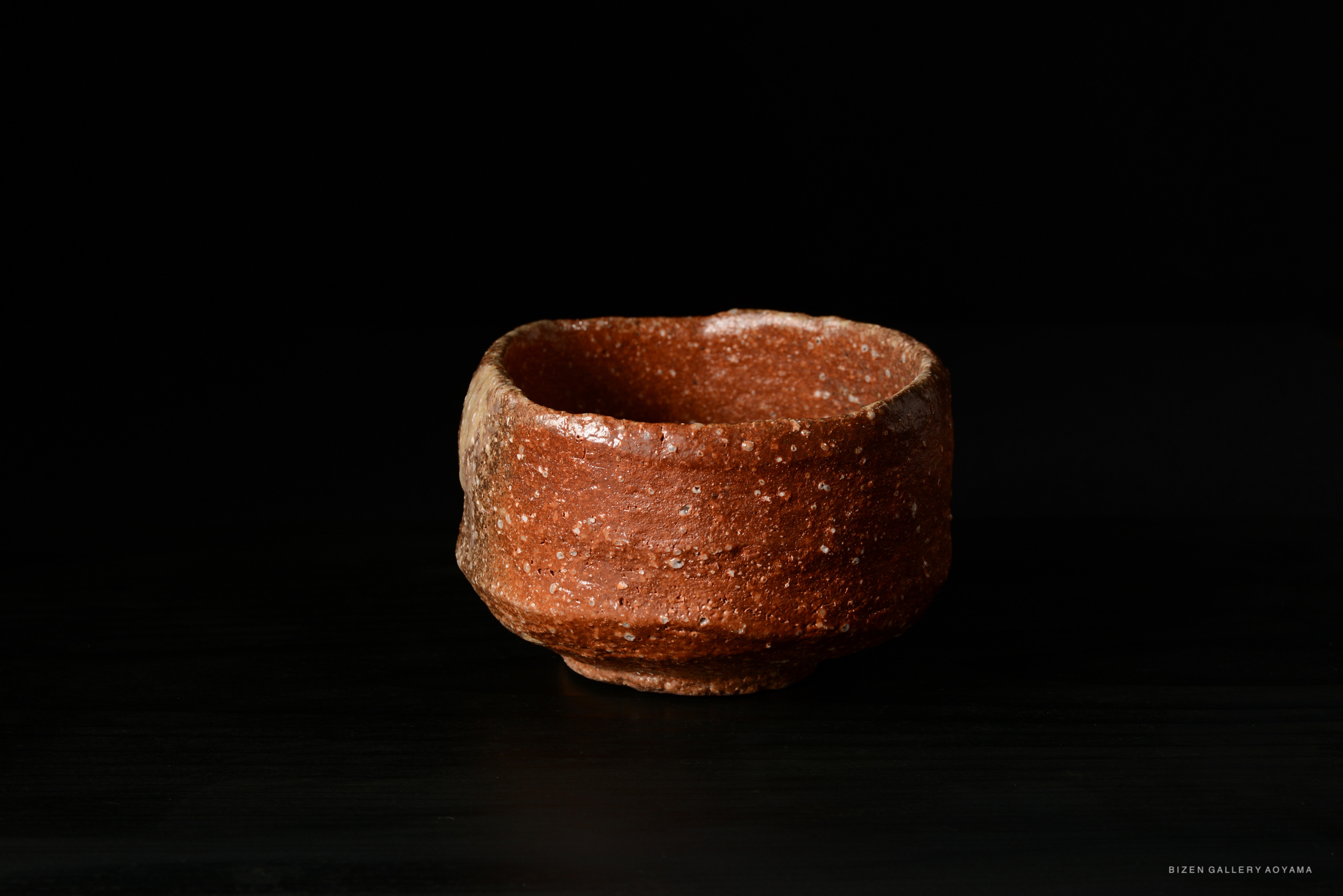 A close-up of a rustic, reddish-brown ceramic bowl with a textured surface, set against a black background.