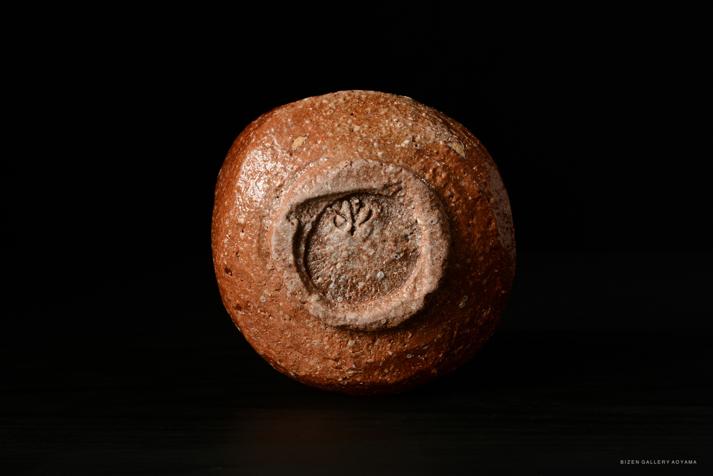 A close-up of a round, textured ceramic piece with a carved design on its surface, set against a dark background.