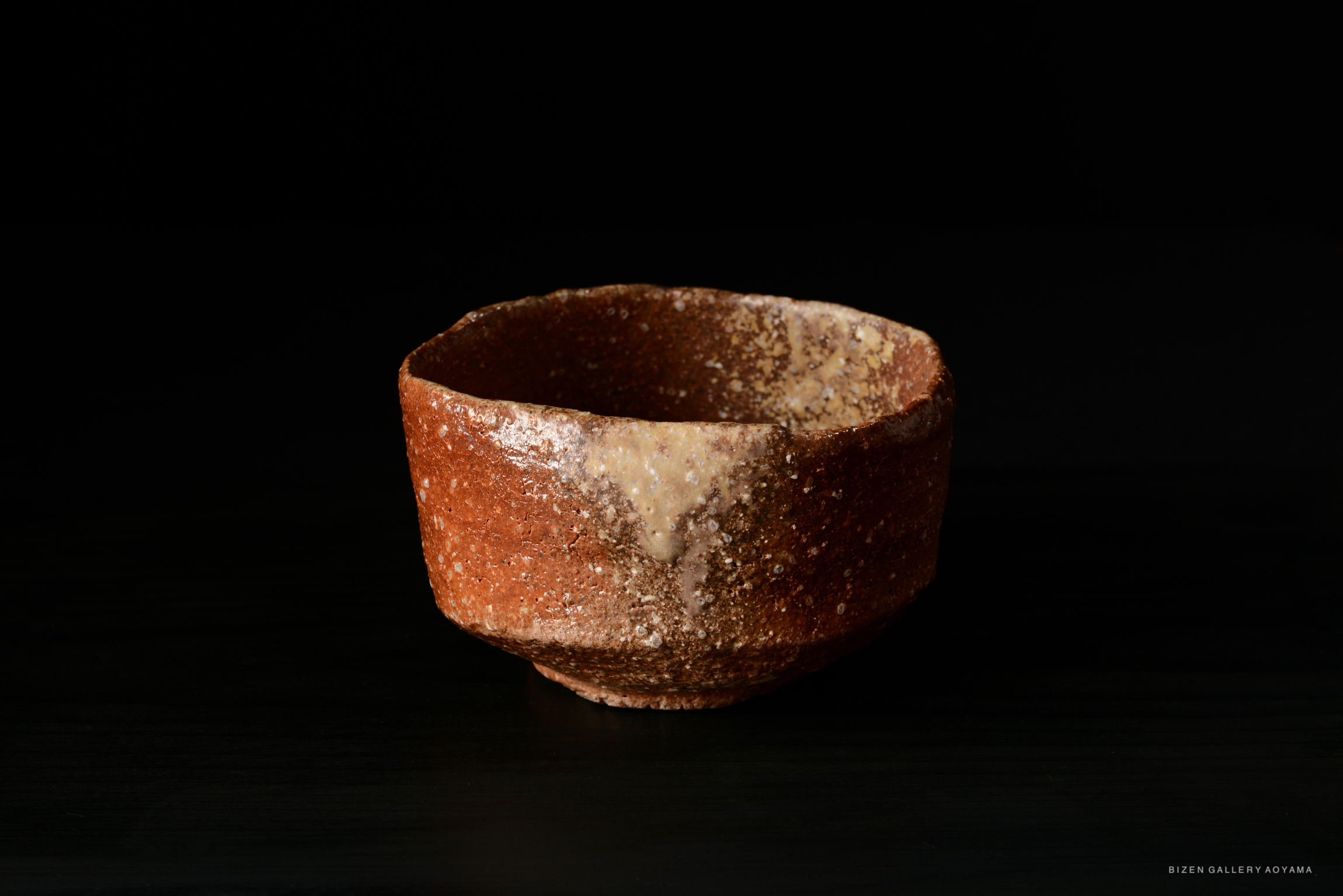 A close-up view of a rustic, handmade ceramic bowl with a textured surface and earthy tones, set against a dark background.