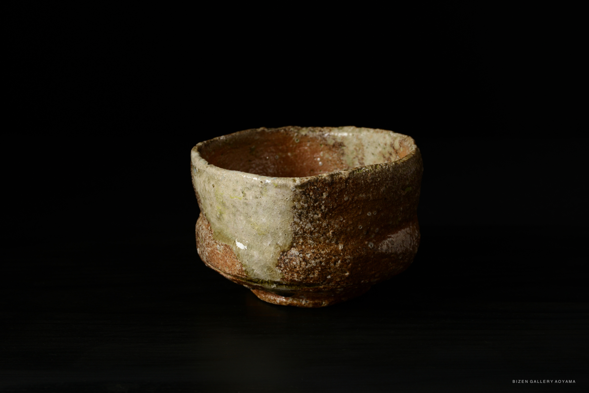 A rustic Shigaraki tea bowl with a textured surface, showcasing earthy tones and a unique shape, set against a dark background.
