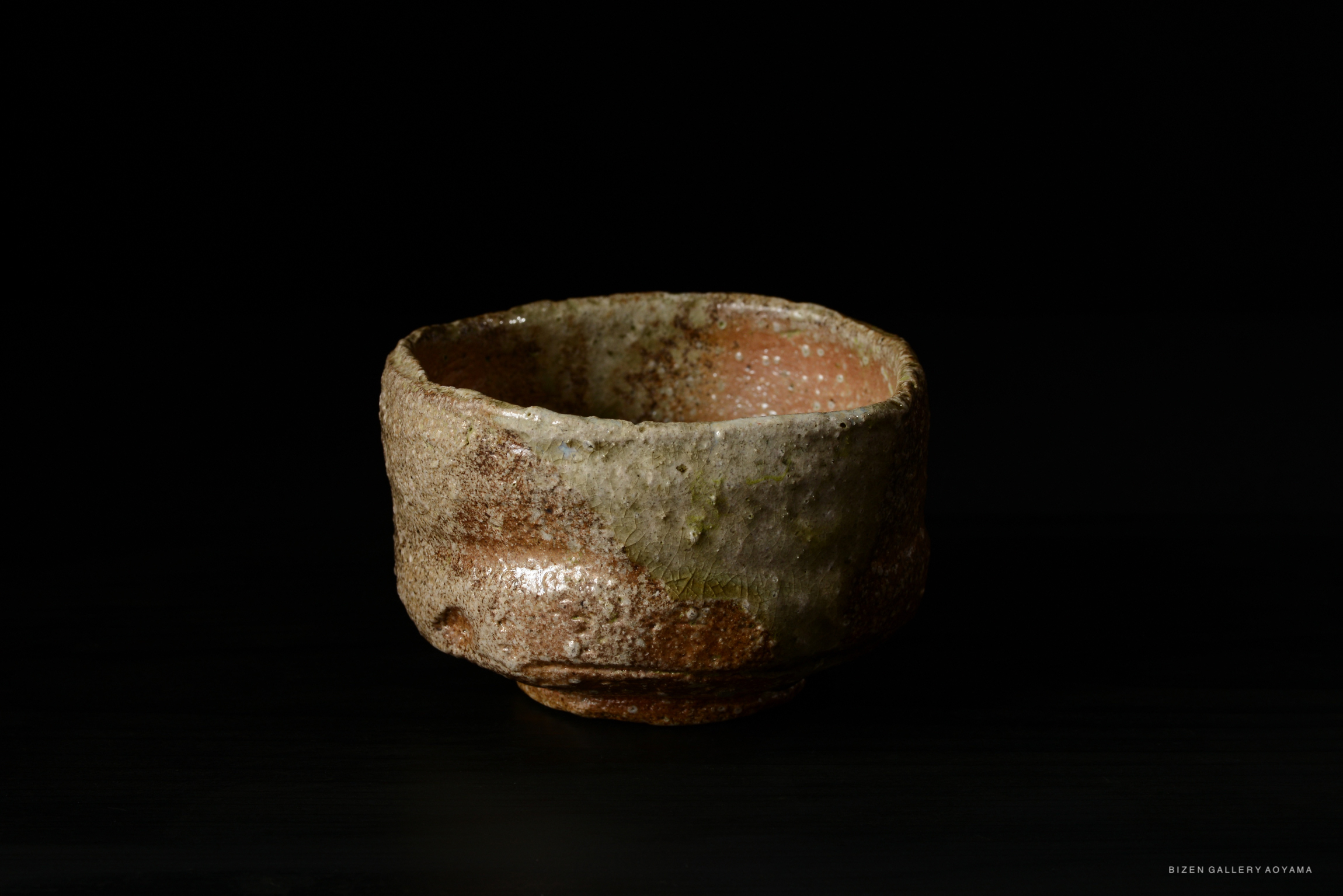 A rustic Shigaraki tea bowl with a textured, earthy surface, showcasing hues of brown and green against a dark background.