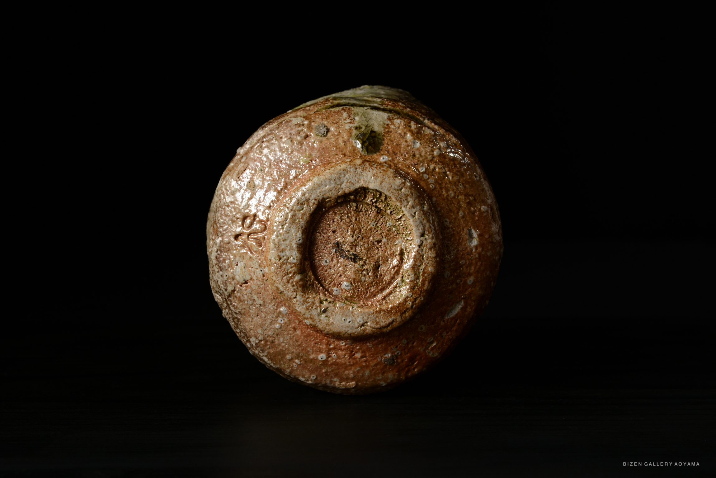 A close-up view of the bottom of a rustic ceramic tea bowl with an earthy texture and muted colors, set against a dark background.