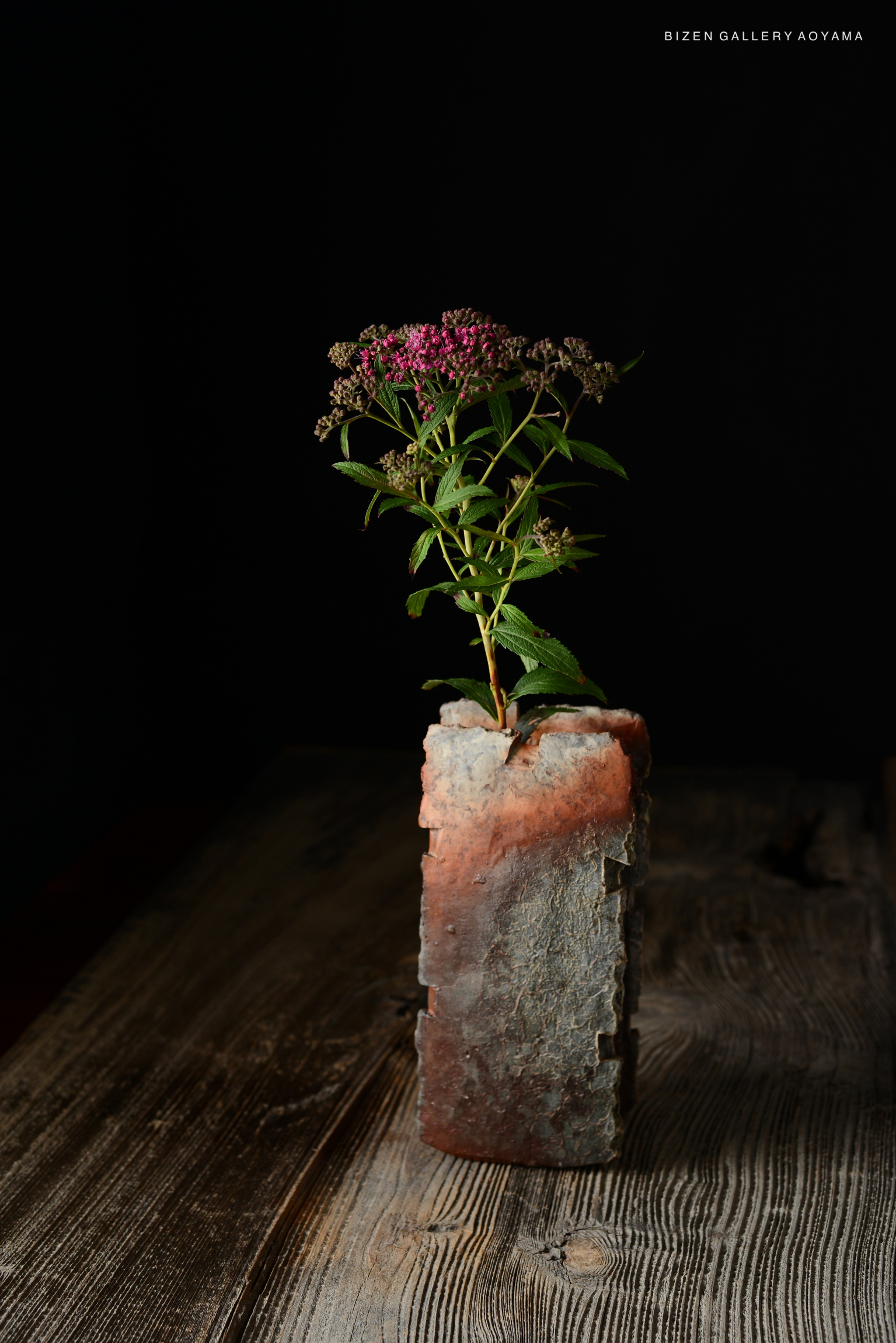 A minimalist ceramic flower vase displayed on a wooden surface, holding a small bouquet of pink flowers. The background is dark, emphasizing the vase's textures and colors.