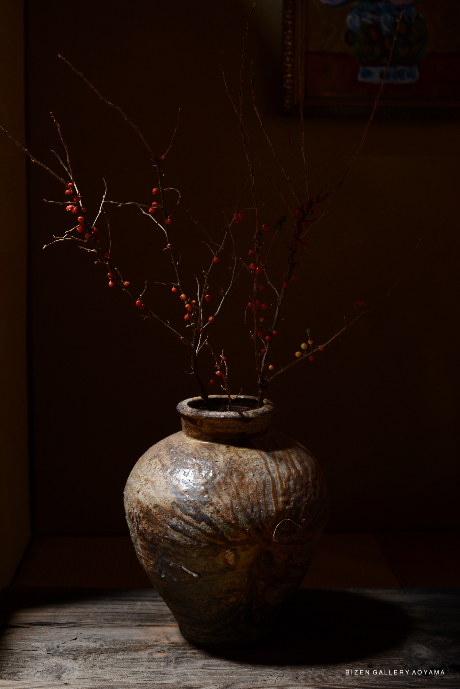 A Bizen pottery vase with decorative branches and red berries, illuminated artistically in a dimly lit space.