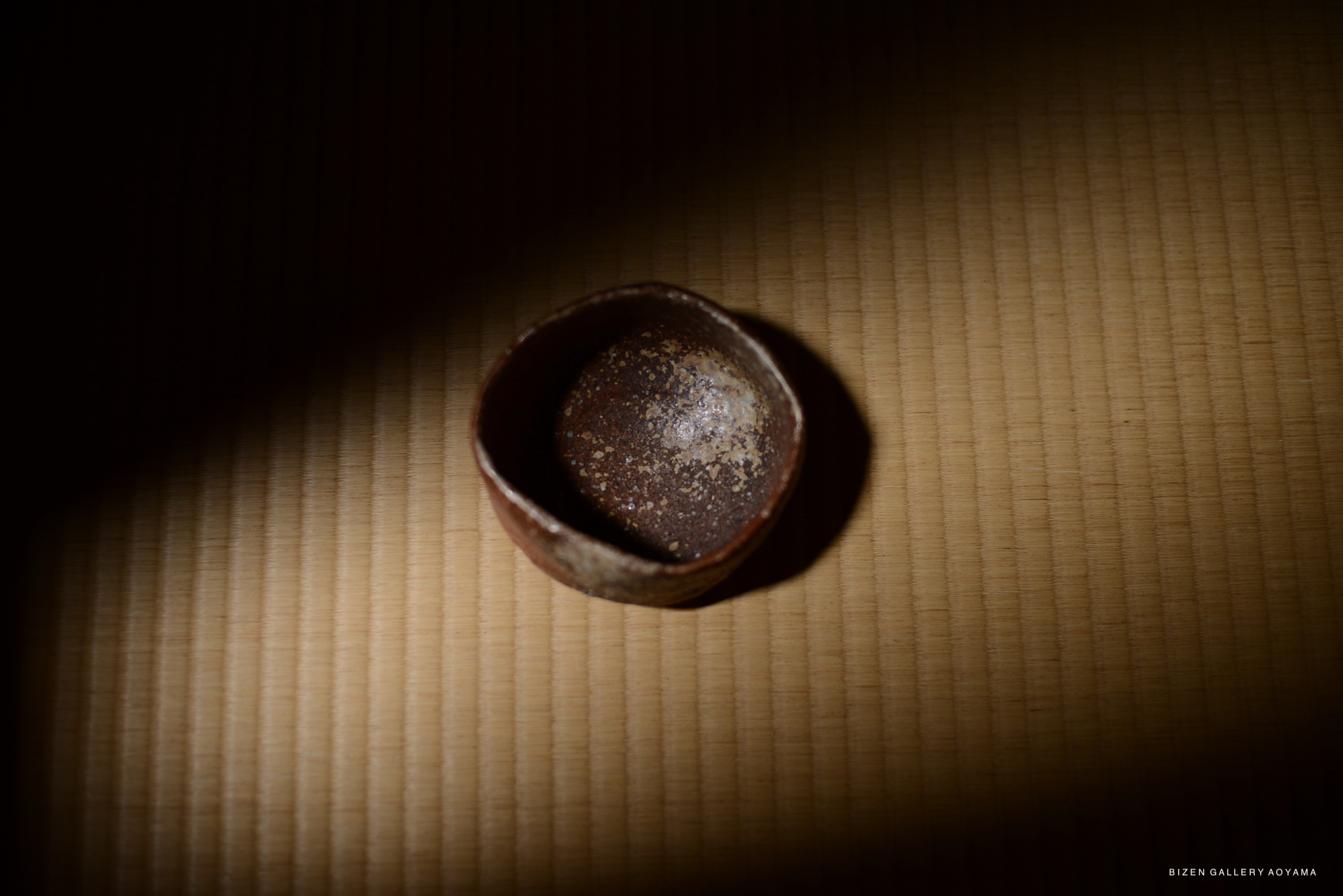 A small, round ceramic dish sitting on a tatami mat, softly illuminated with a spotlight.