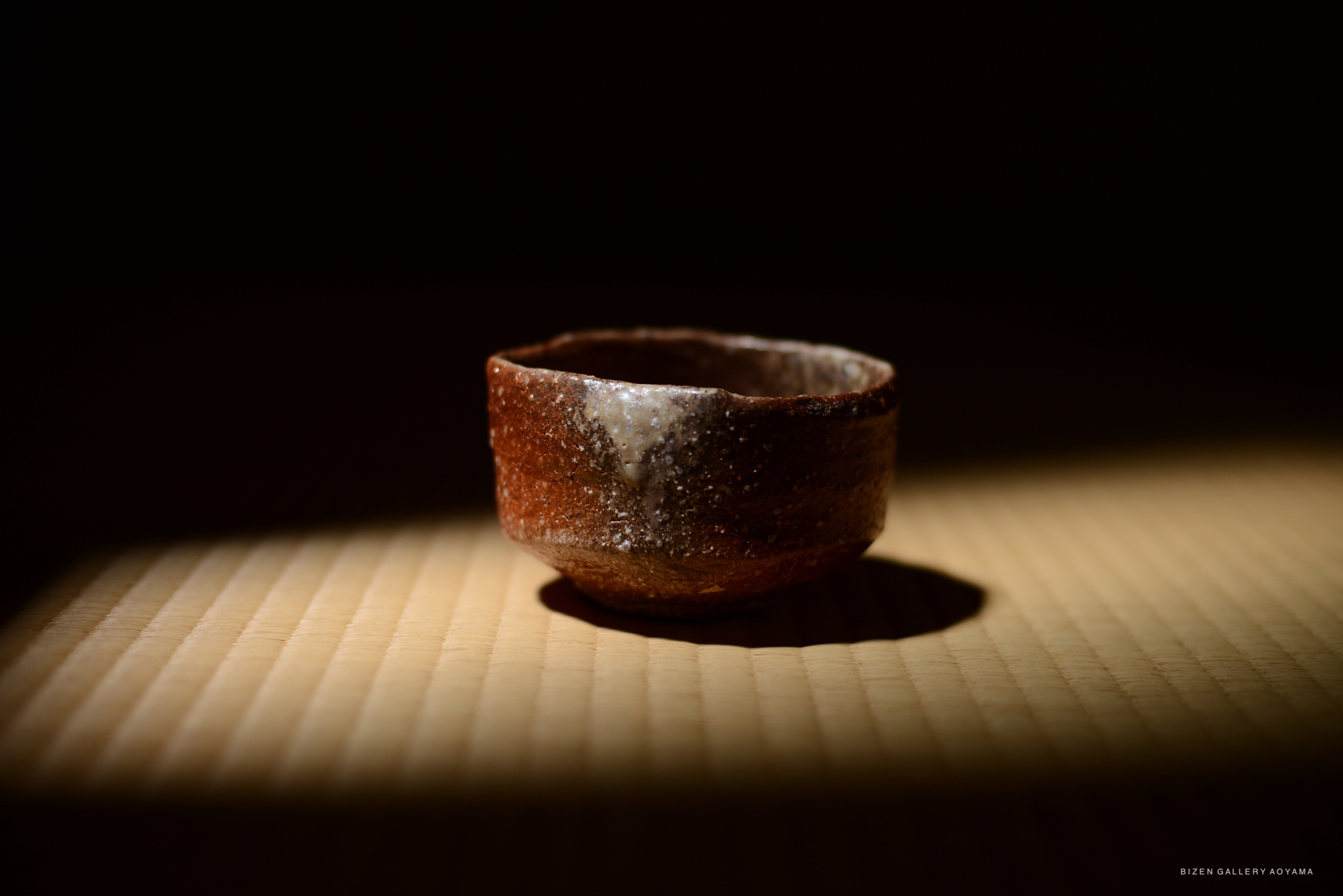 A close-up of a small Shigaraki chawan tea bowl with a textured brown exterior, softly illuminated by light, resting on a tatami mat.