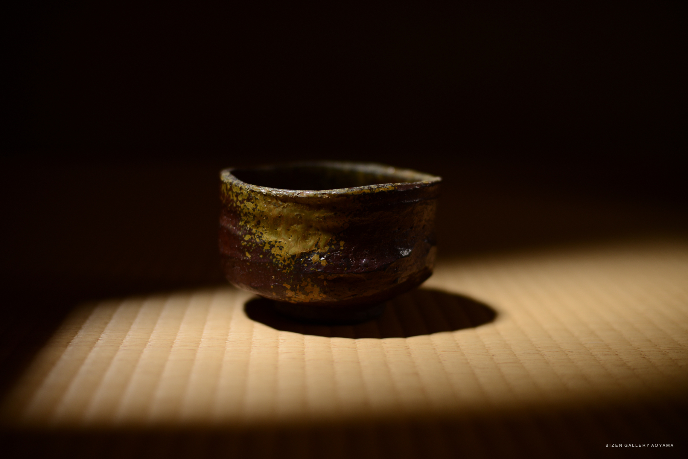 A beautifully crafted Bizen chawan (tea bowl) in warm reddish brown with a textured surface, illuminated softly on tatami mats.
